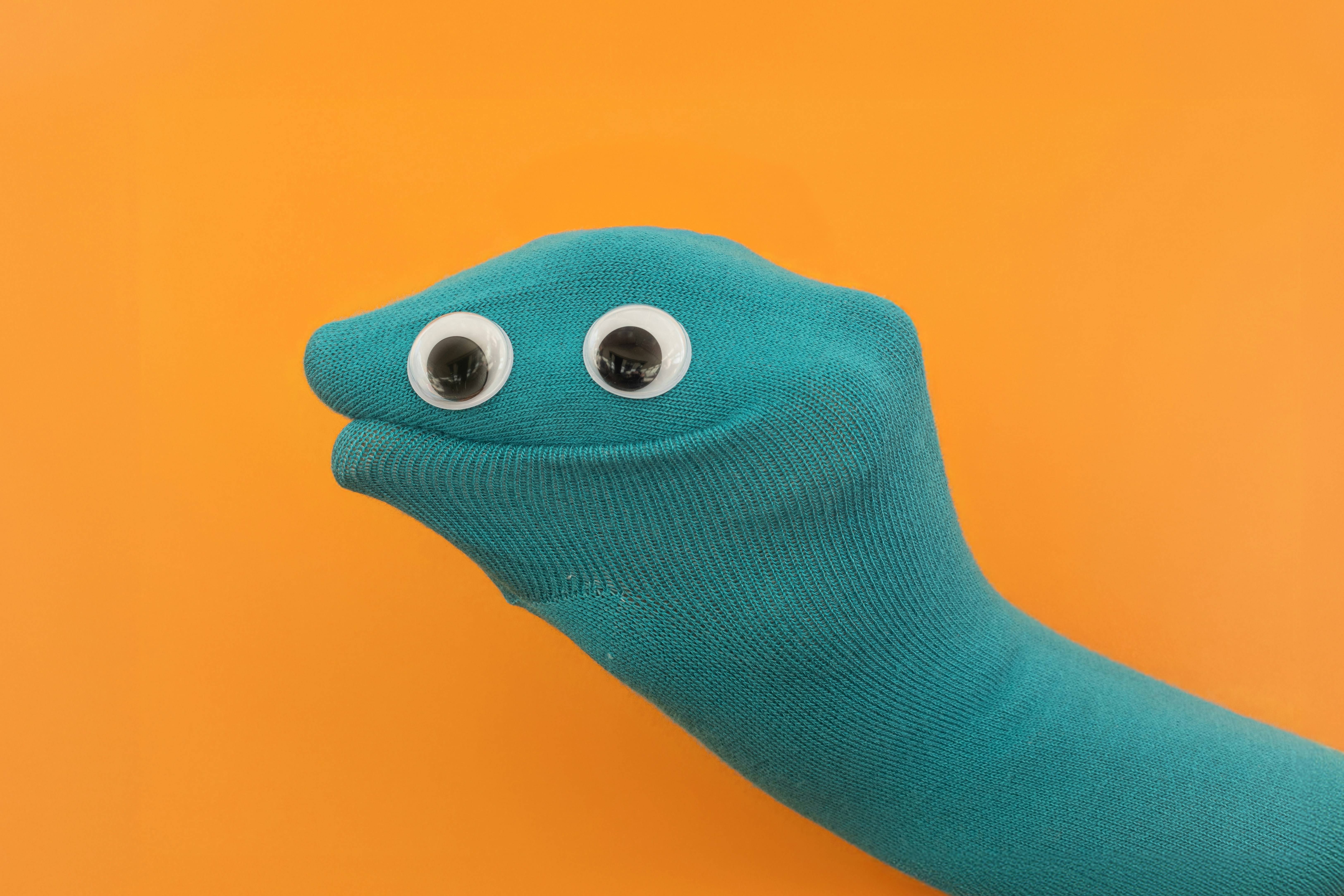 A smiling, jade coloured, sock puppet with googly eyes set against an orange background.