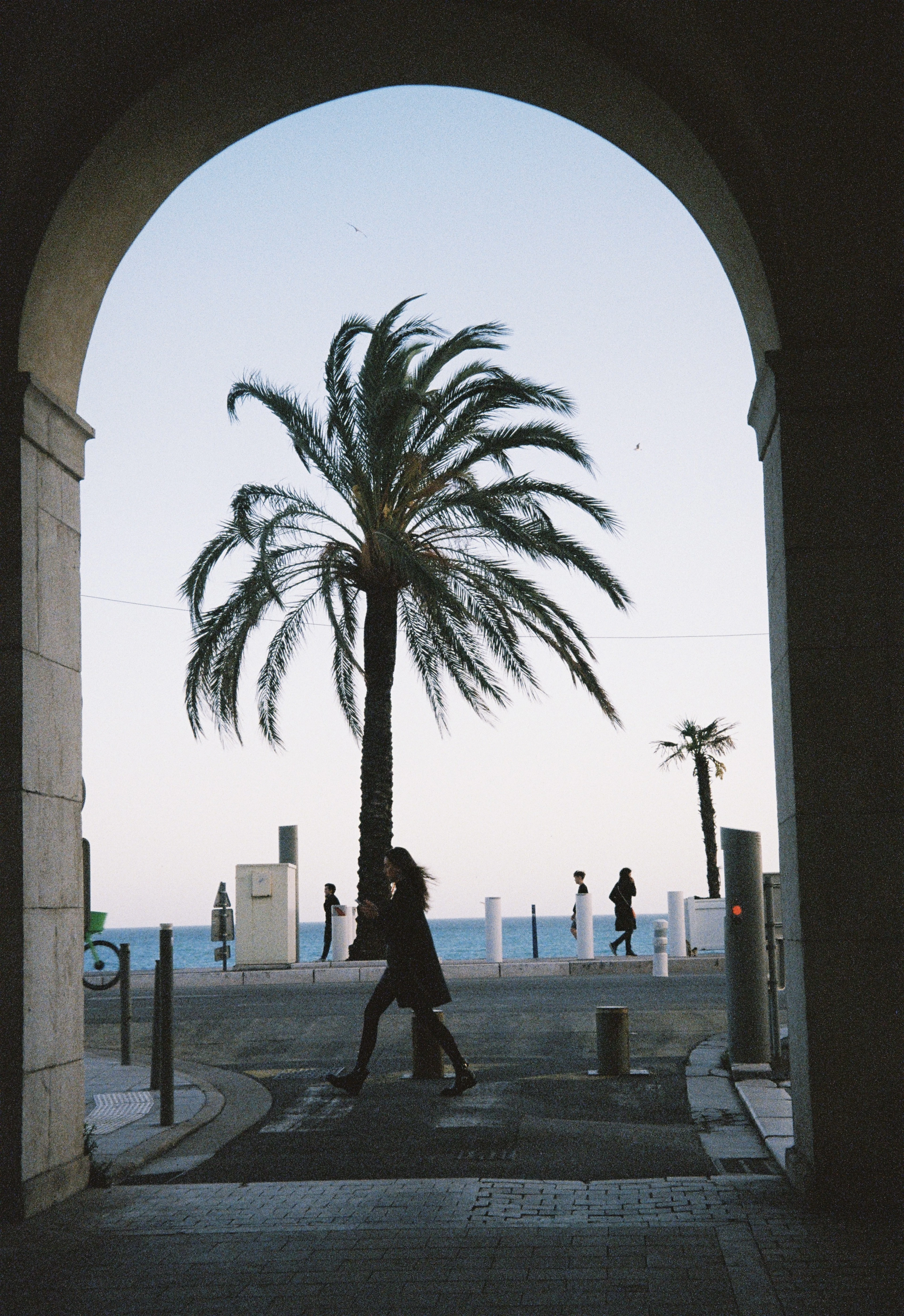 A silhouetted pedestrian walks beneath a stone archway framing a palm tree and the sea at dusk in Nice.