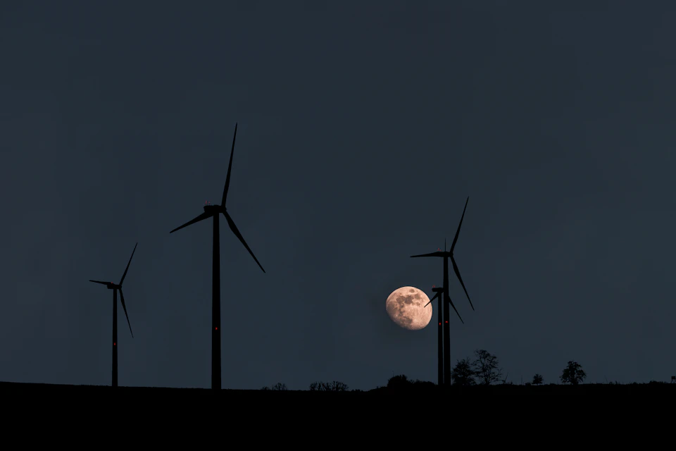 The image depicts a serene night scene with three wind turbines standing tall against the dark sky, their blades spinning silently. The largest turbine is positioned on the left side and the other two are located more towards the center of the frame. In the background, a full moon can be seen in the top right corner, casting a soft glow over the landscape below.