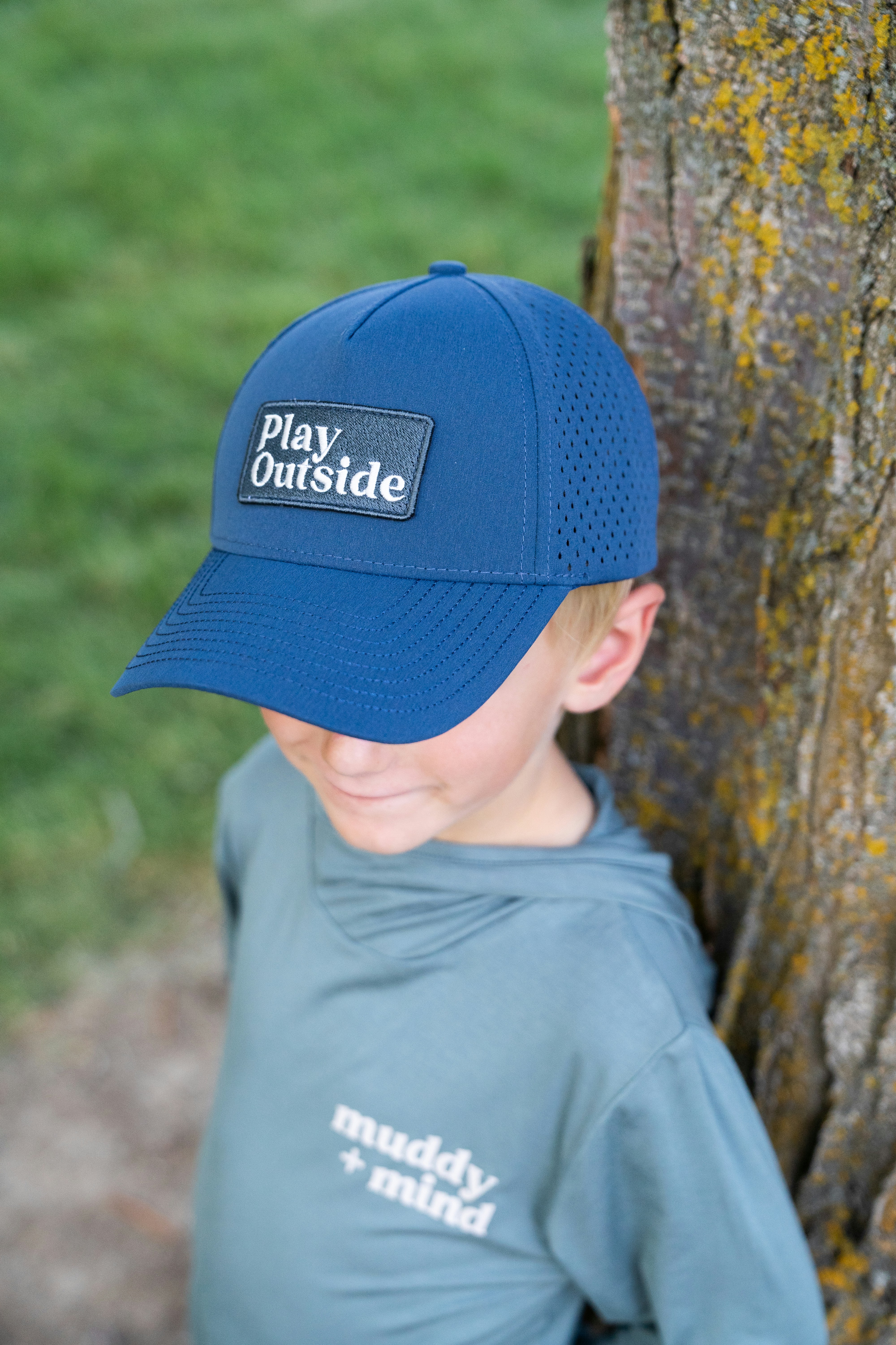 A young boy wearing a blue hat standing next to a tree