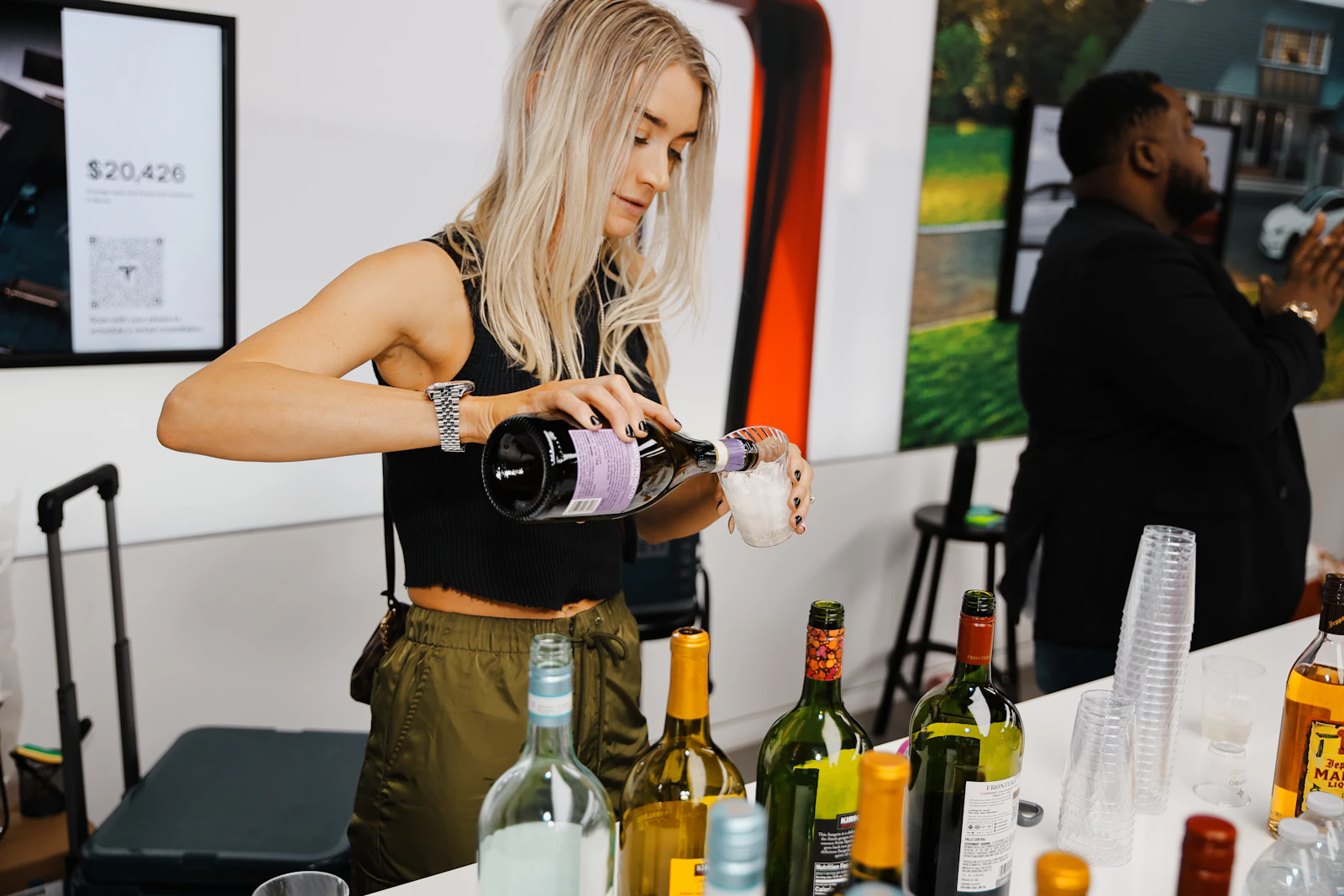 A woman standing behind a table of bottles at an event, serving a drink to someone off camera