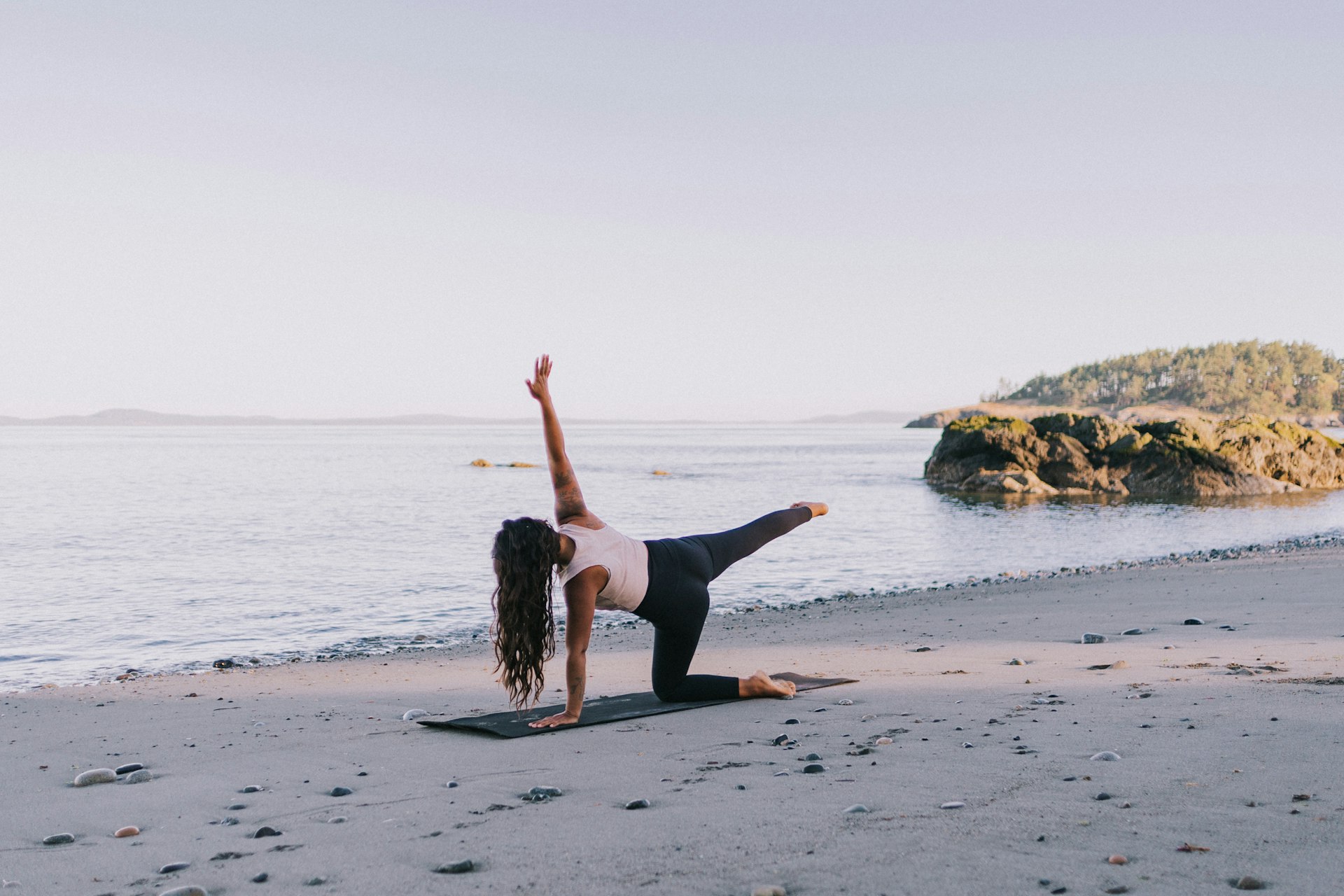 Woman on a yoga mat on a beach in a kneeling half moon variation, one knee and one hand on the mat, the other leg and arm lifted