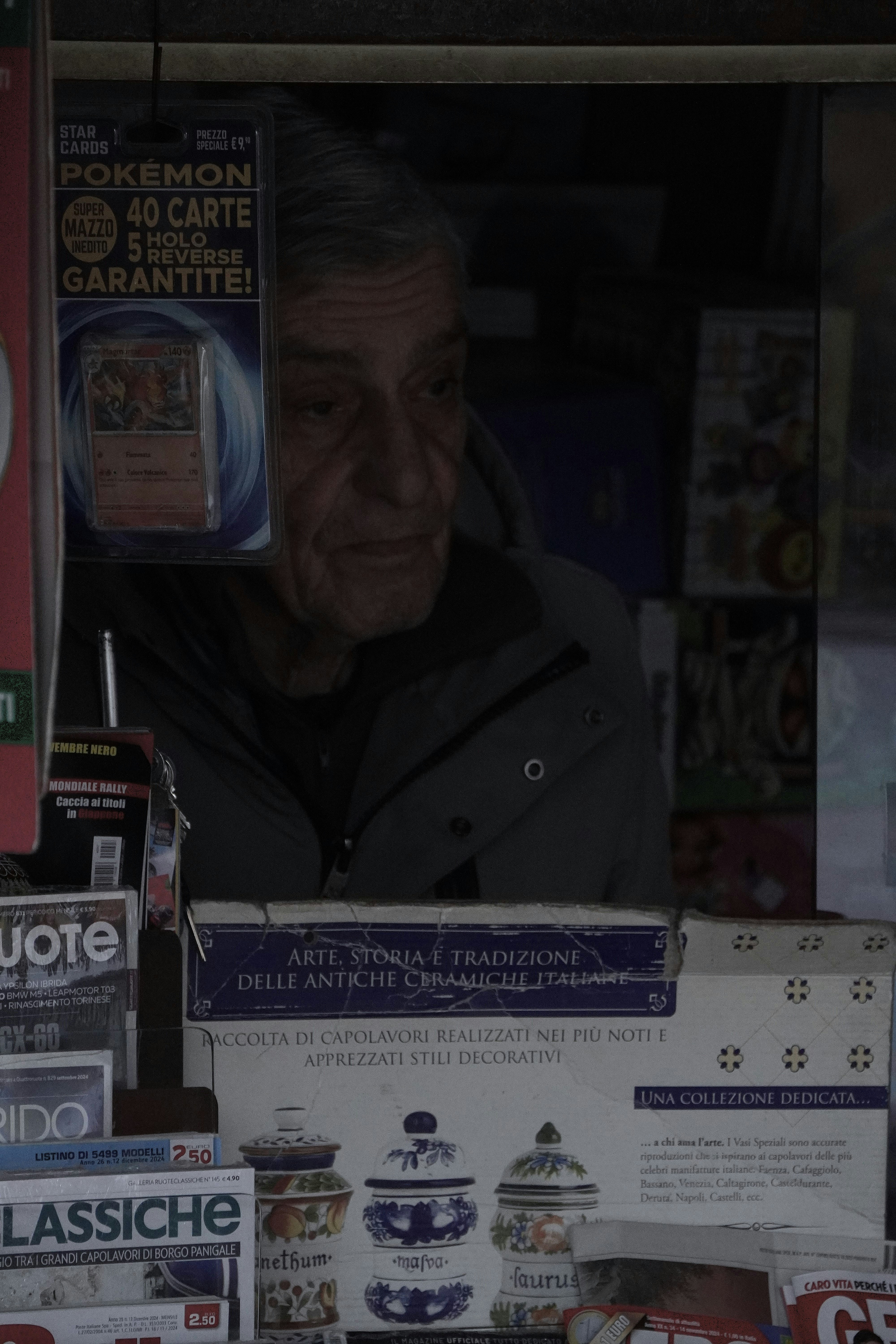 An elderly newsagent, with a tired expression, is talking to a rather demanding lady