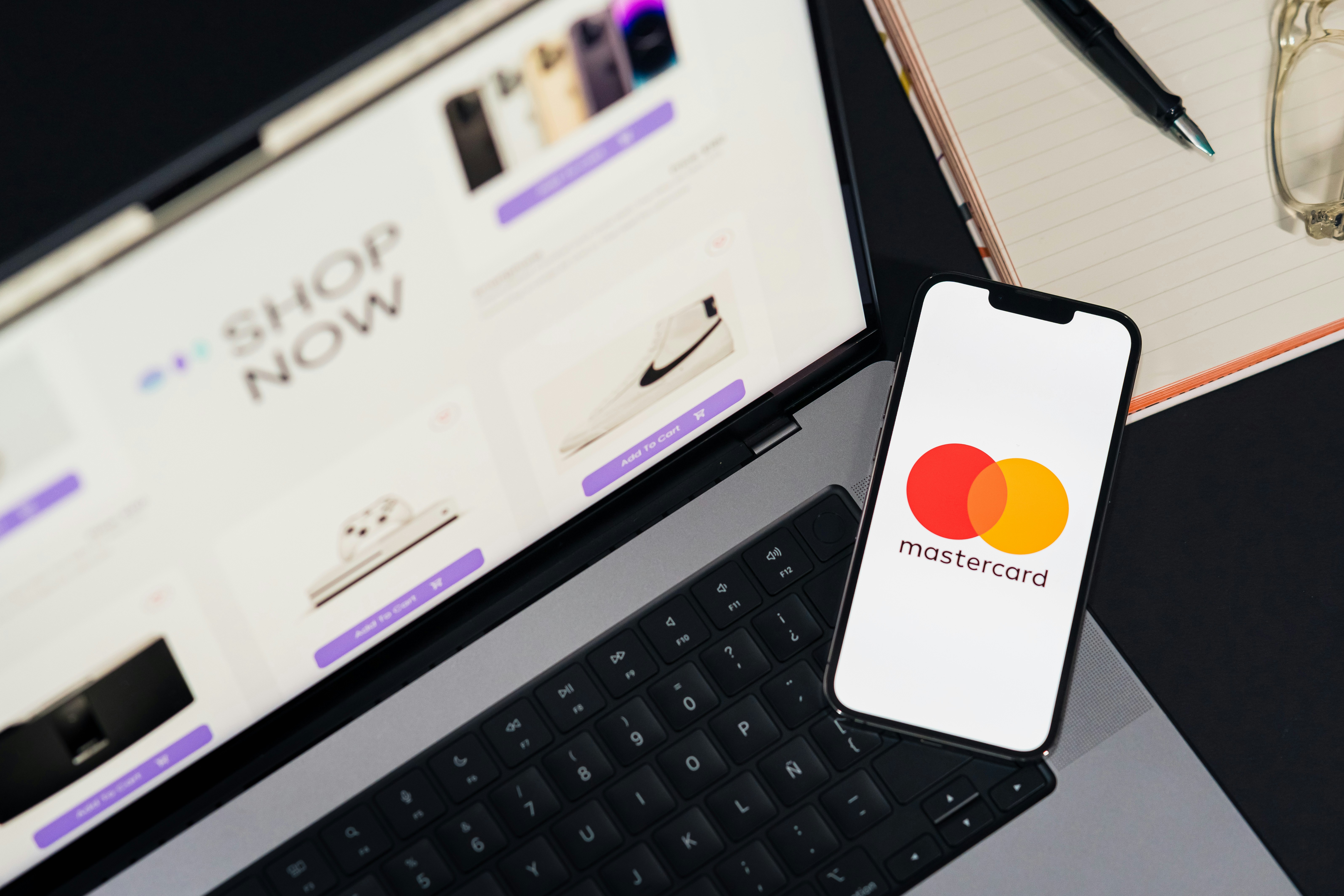 Mastercard logo on phone screen. Top view of workspace. Table desk with laptop with ecommerce on screen, mobile, notepad