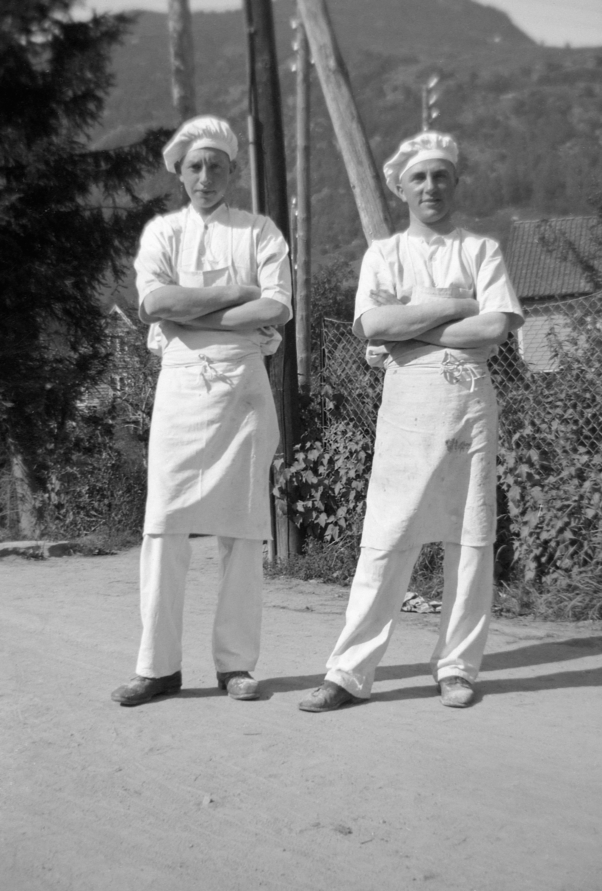 Two bakers in uniform standing outdoors.