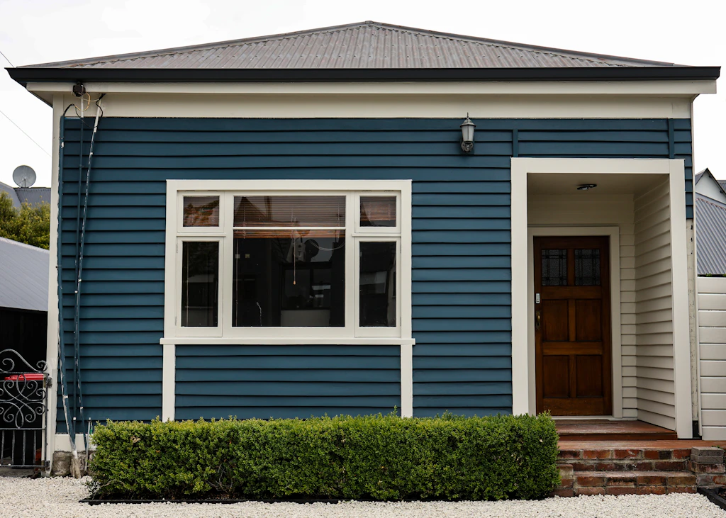 Blue weatherboard bungalow with white window trim, a timber front door and a neat hedge