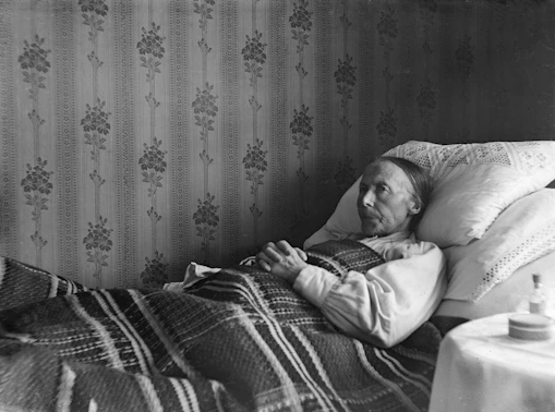An elderly person lies in bed, covered by a blanket.