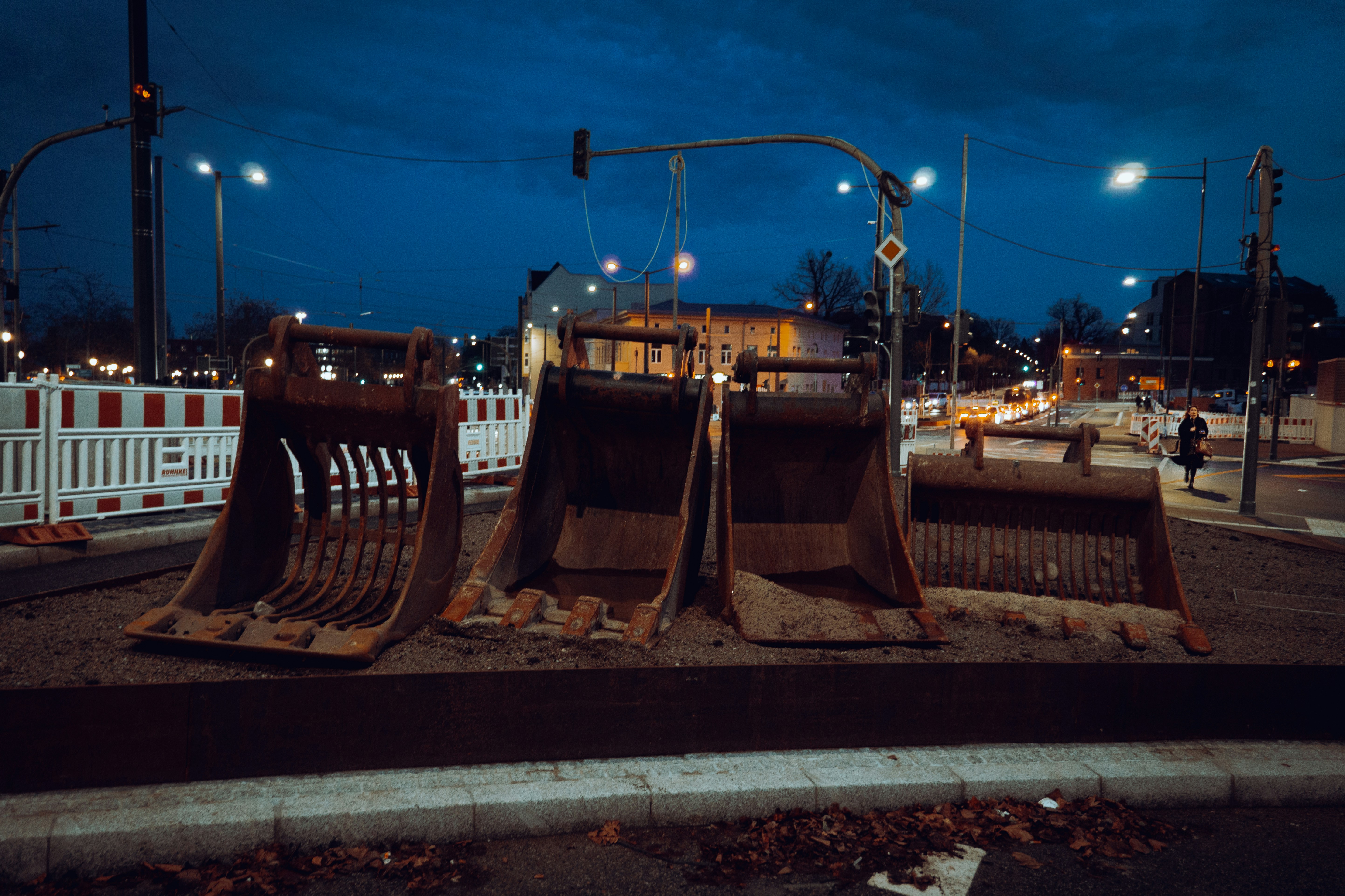 A dimly lit street at night with an open, unguarded construction drain, a few construction materials scattered nearby. The mood is somber and dangerous.