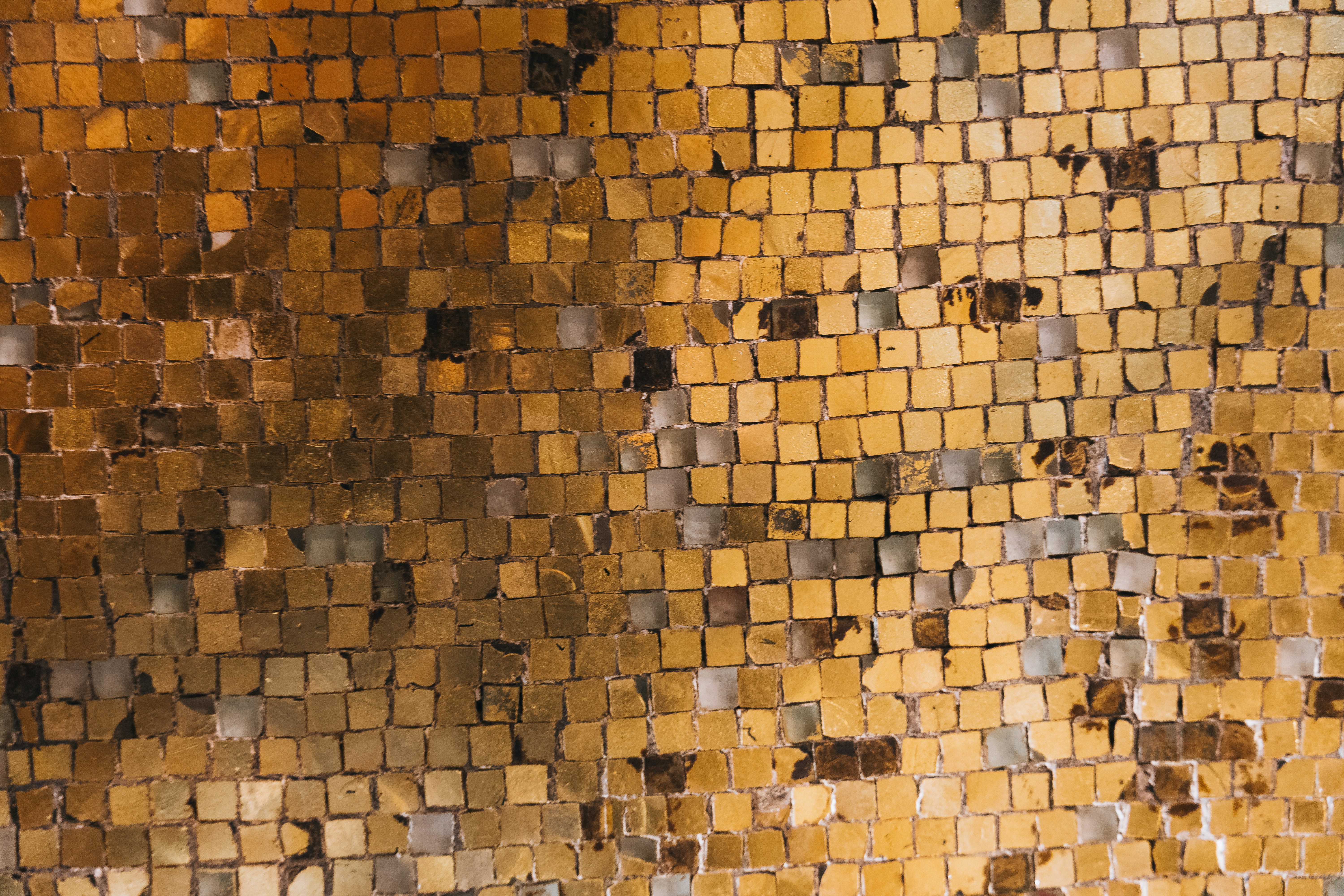A mesmerizing mosaic of golden tiles, each square reflecting light with subtle variations of bronze, amber, and muted metallic tones. The textured surface reveals an intricate blend of earthy warmth and shimmering elegance, evoking a sense of timeless craftsmanship.