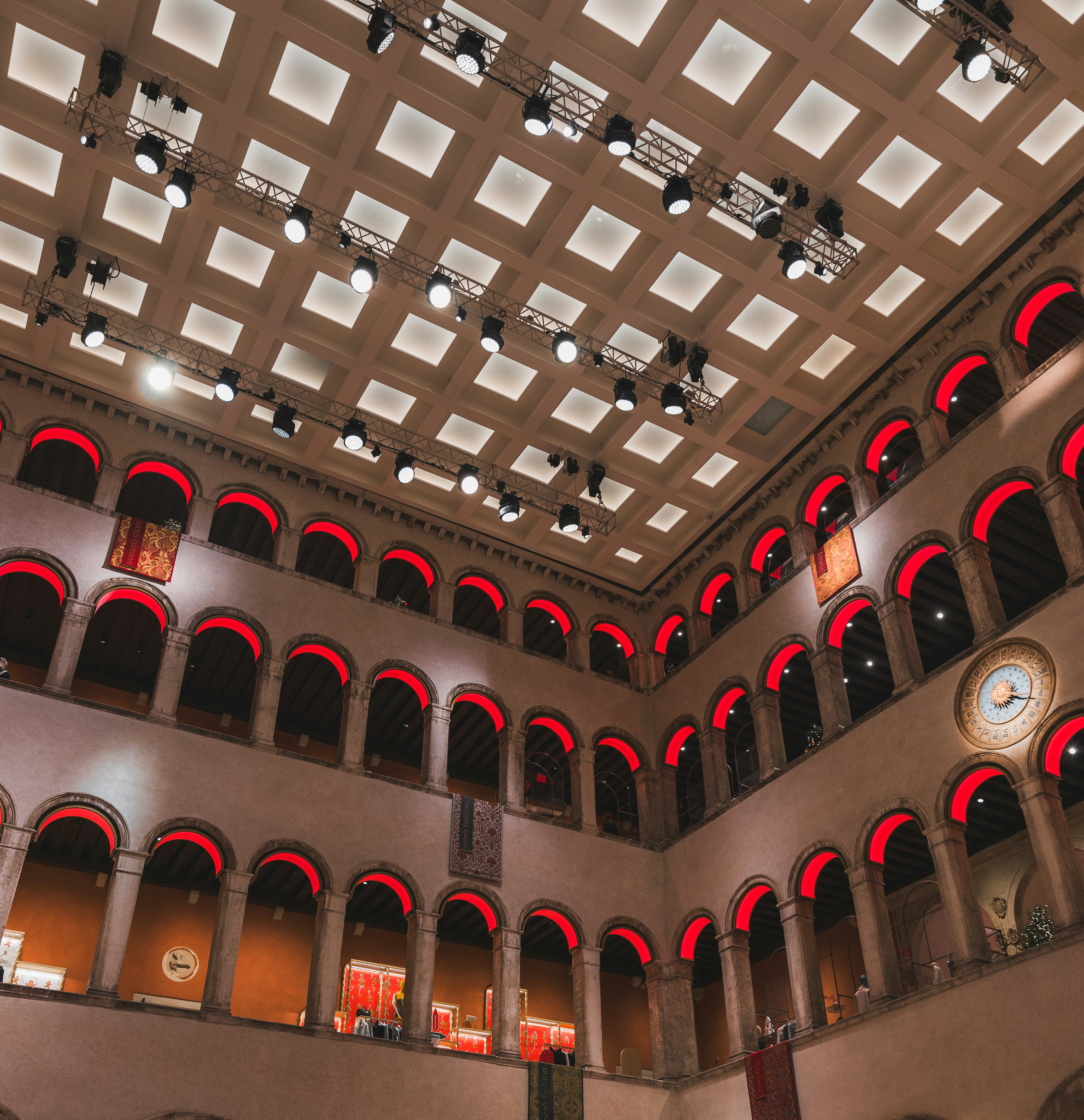 An ornate interior with grand arches glowing in vibrant red light, arranged symmetrically under a grid-like ceiling illuminated by modern spotlights. A fusion of historical elegance and contemporary design creates a striking visual harmony.