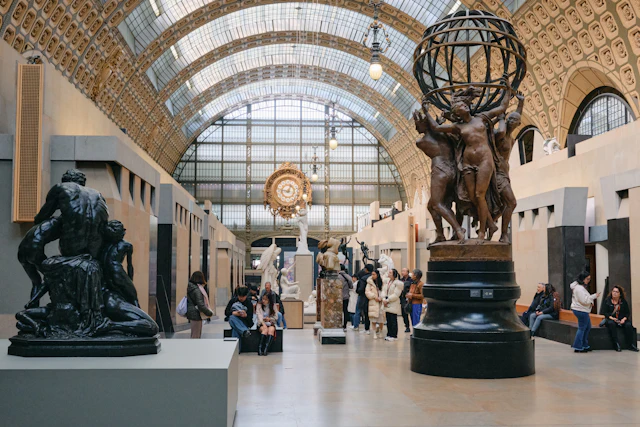 The grand nave of the Musée d'Orsay under its glass barrel-vault ceiling, with the gilded station clock and the bronze globe sculpture among visitors.