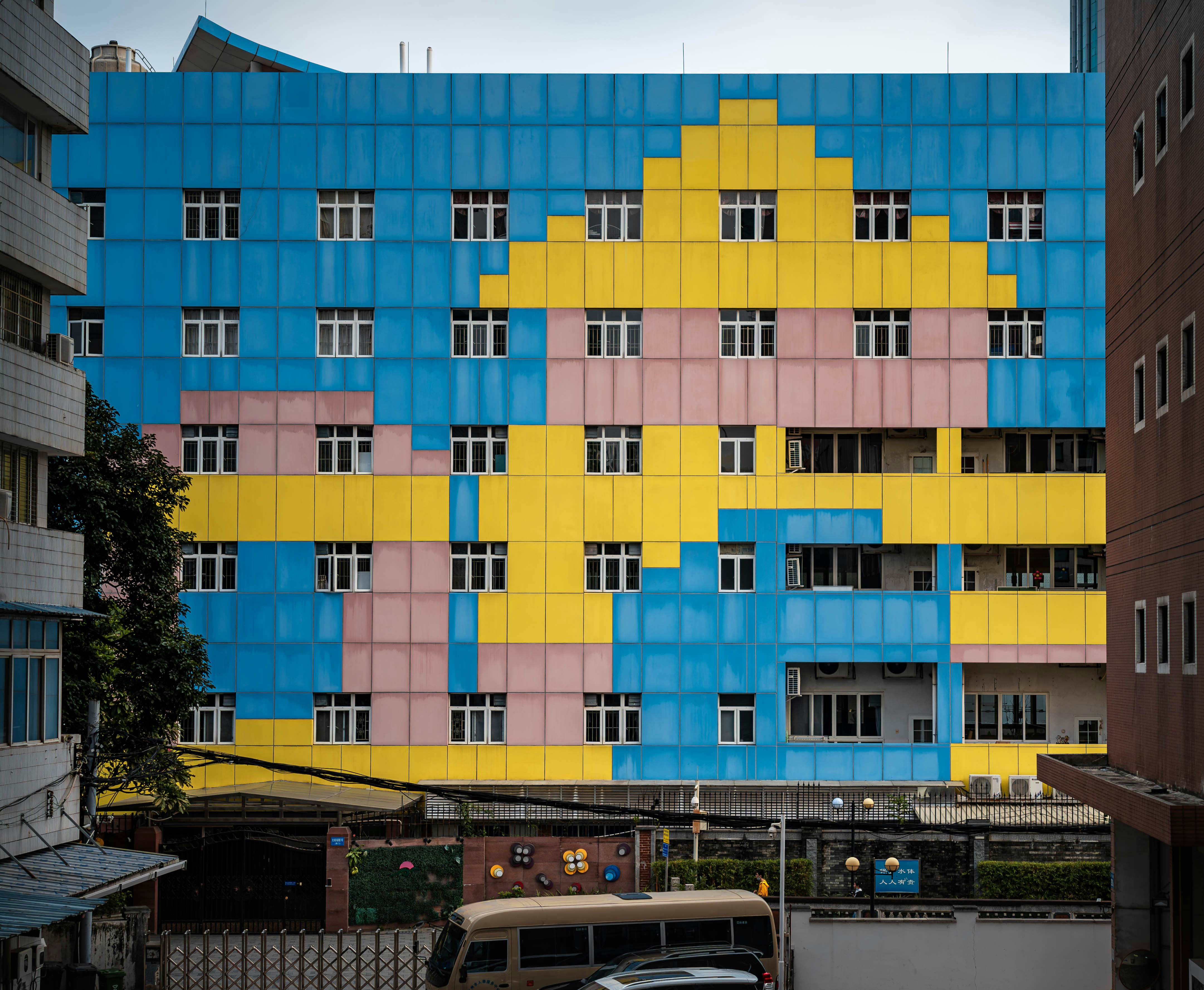 The facade of a multi-story building presents a striking visual with its bold color block design. Large panels in vibrant blue, yellow, and pastel pink are arranged in a geometric, almost pixelated pattern across the exterior, punctuated by numerous, regularly spaced white windows. Flanked by parts of adjacent buildings, this colorful architecture stands out in the urban environment.