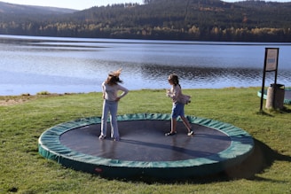 Two young girls standing on a trampoline near a lake