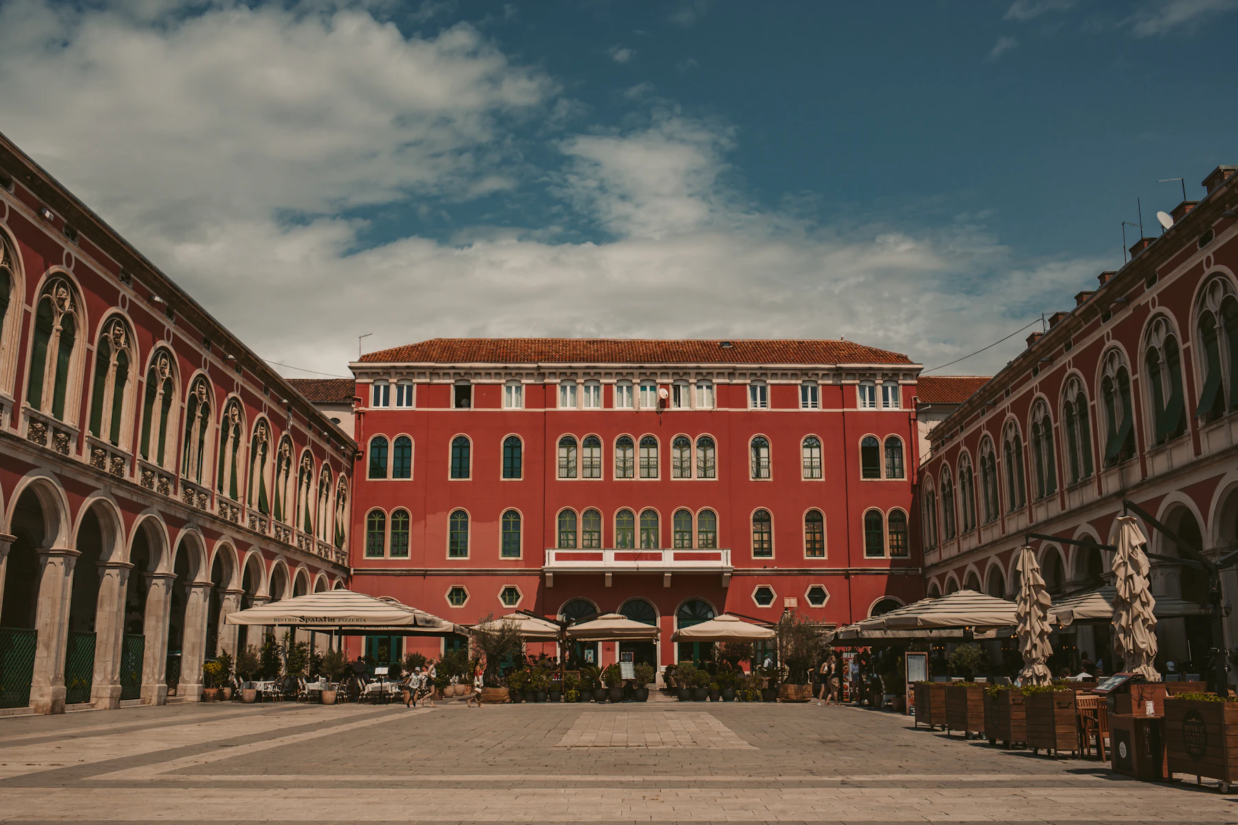 Tables and umbrellas sit in a paved courtyard by a red building.