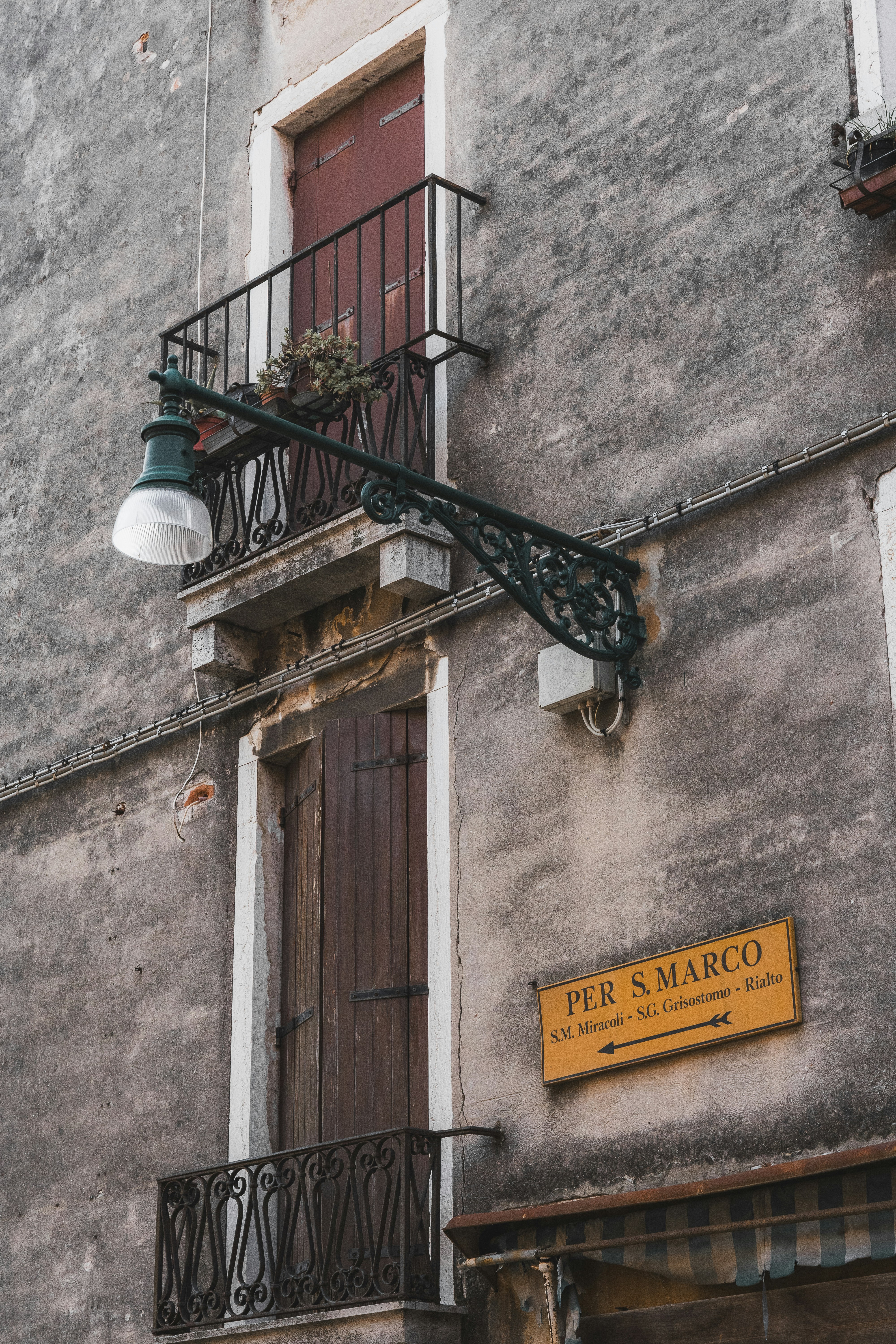A weathered stone facade featuring a rustic wooden door flanked by wrought-iron balconies, adorned with potted plants. A vintage streetlamp and a yellow directional sign reading "Per S. Marco" add charm and guide viewers through the timeless streets of Venice.