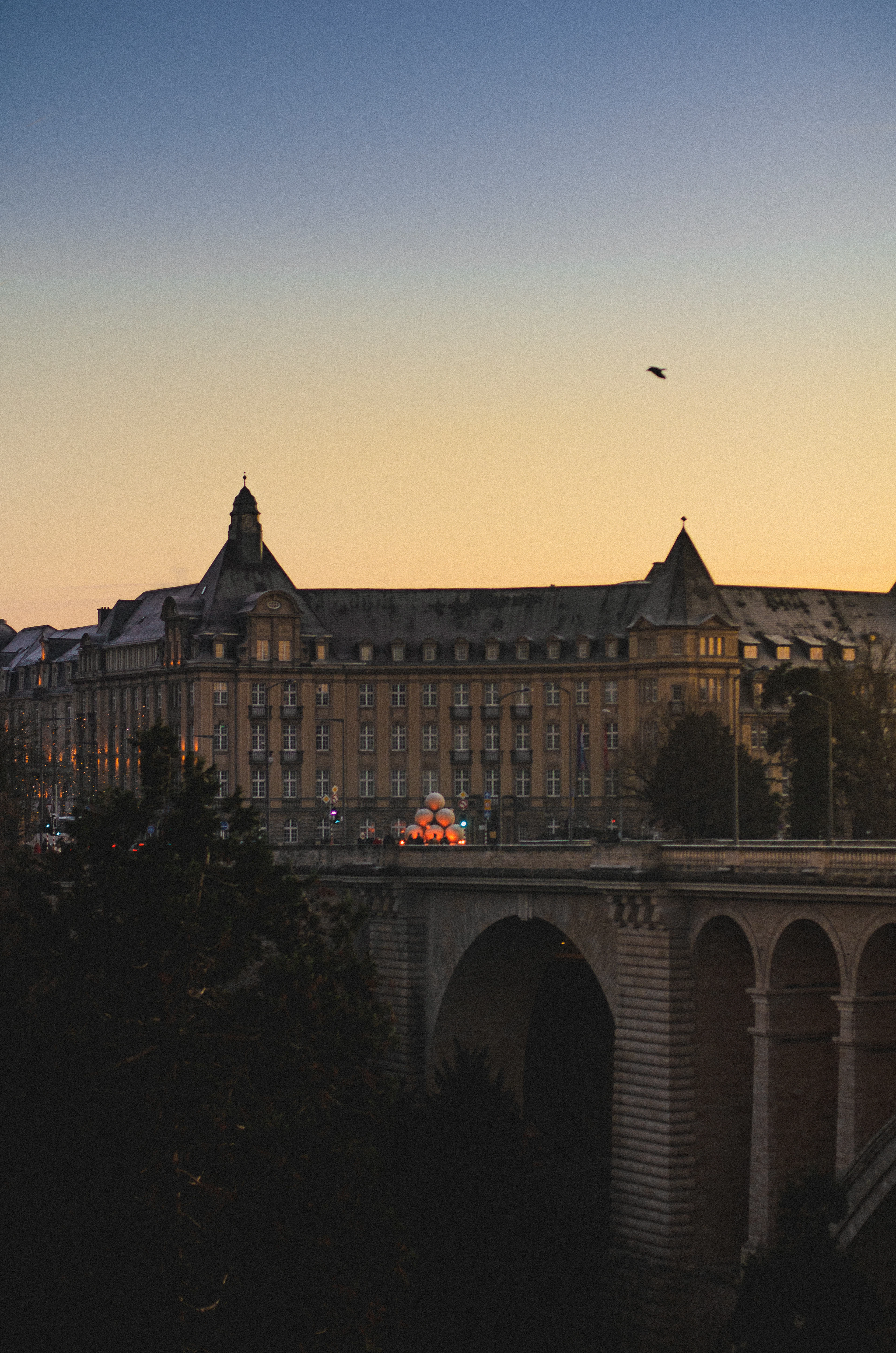 A stone arch bridge crosses a valley at dusk, with an ornate building and a bird in flight against the sunset sky.