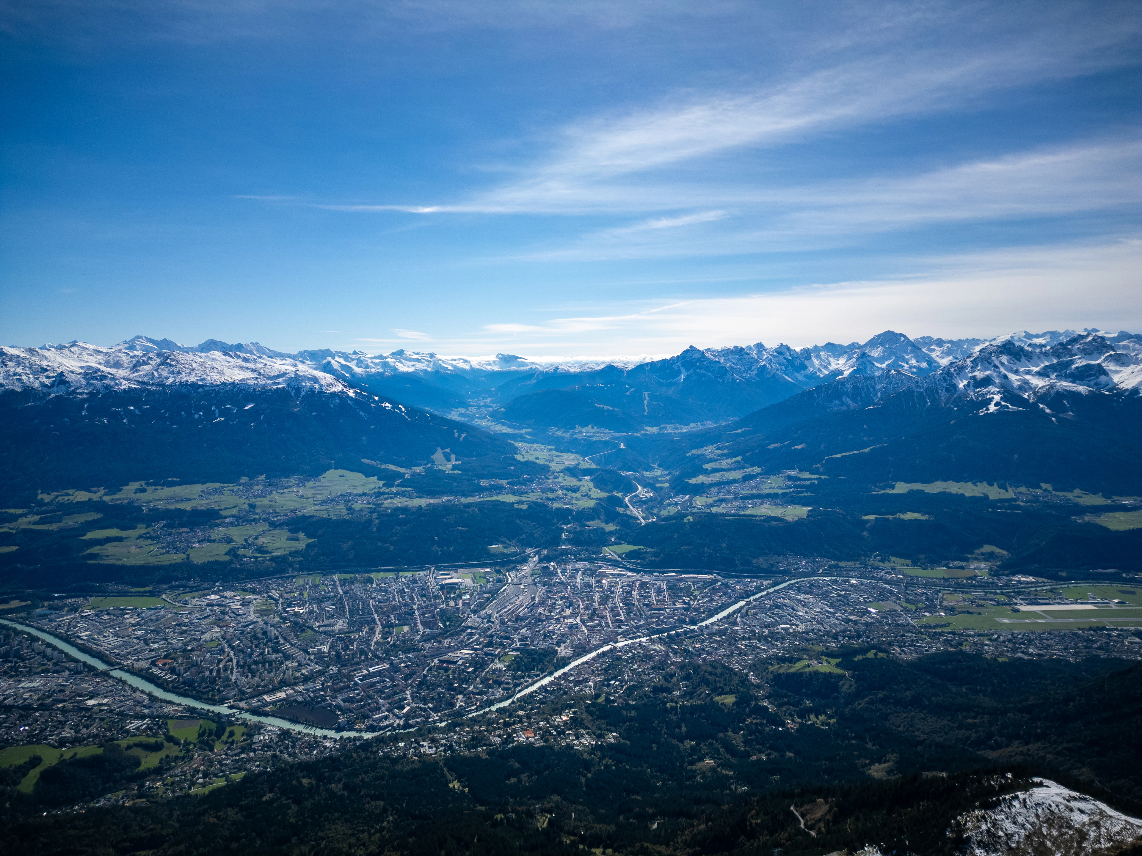 Innsbruck seen from high above, the whole city filling the floor of the Inn valley with the pale river curving through it and range after range of snow-capped Alps stretching away under a deep blue sky