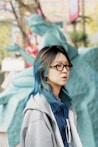 A woman with blue hair and glasses standing in front of a fountain
