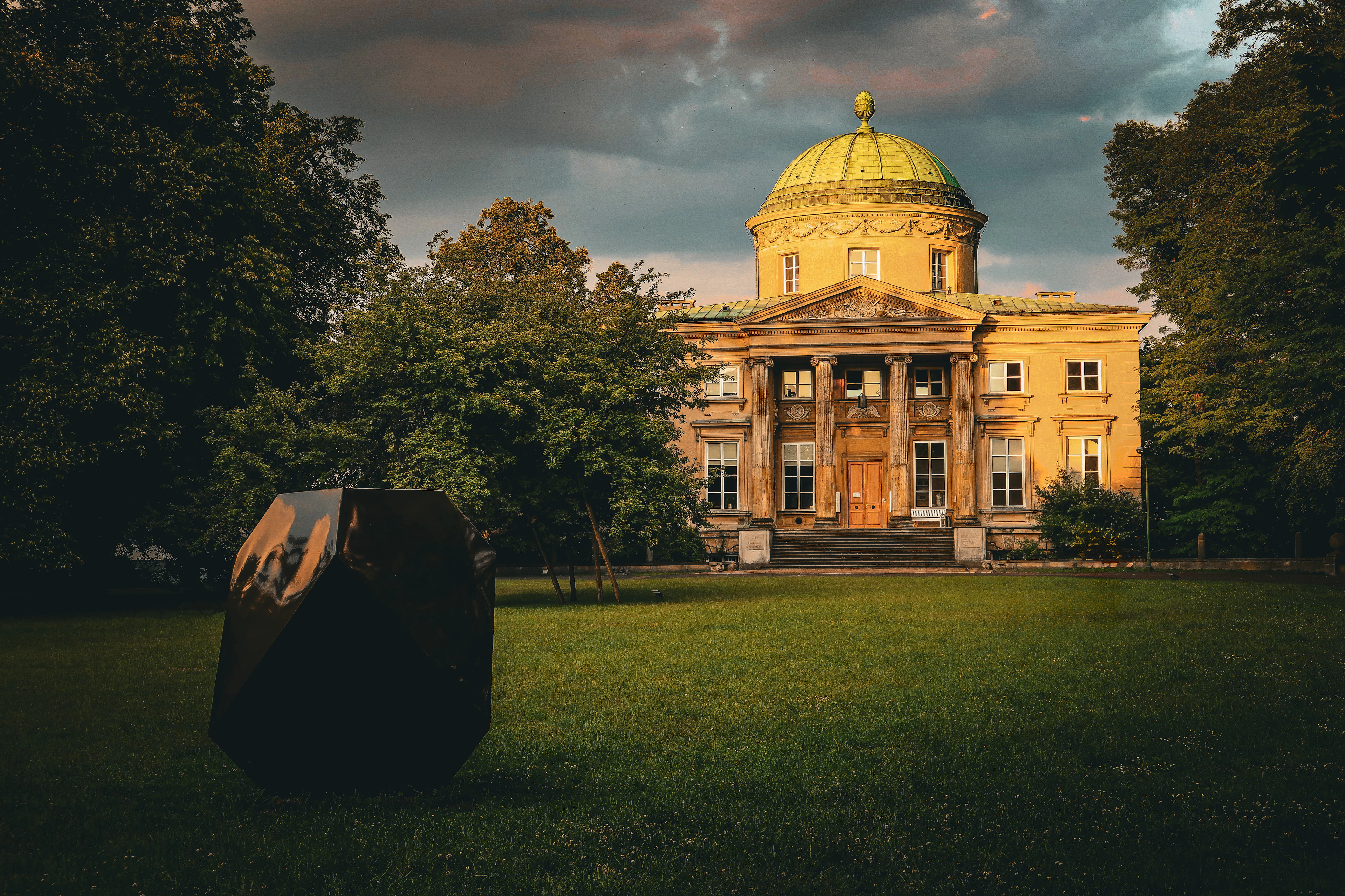 A domed neoclassical palace glows gold at sunset behind a large black geometric sculpture on the lawn