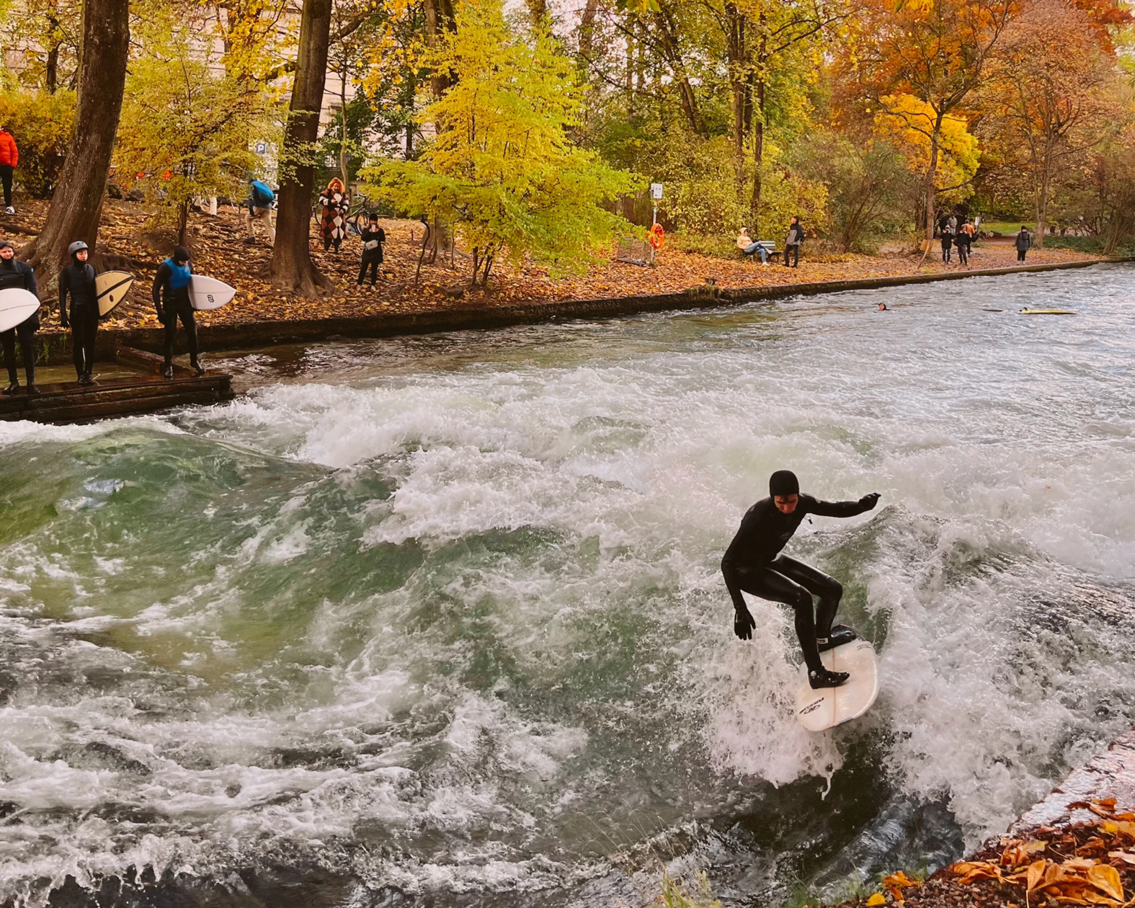 A wetsuited surfer riding the standing wave on the Eisbach river channel in Munich's Englischer Garten, with autumn trees and waiting surfers on the bank.