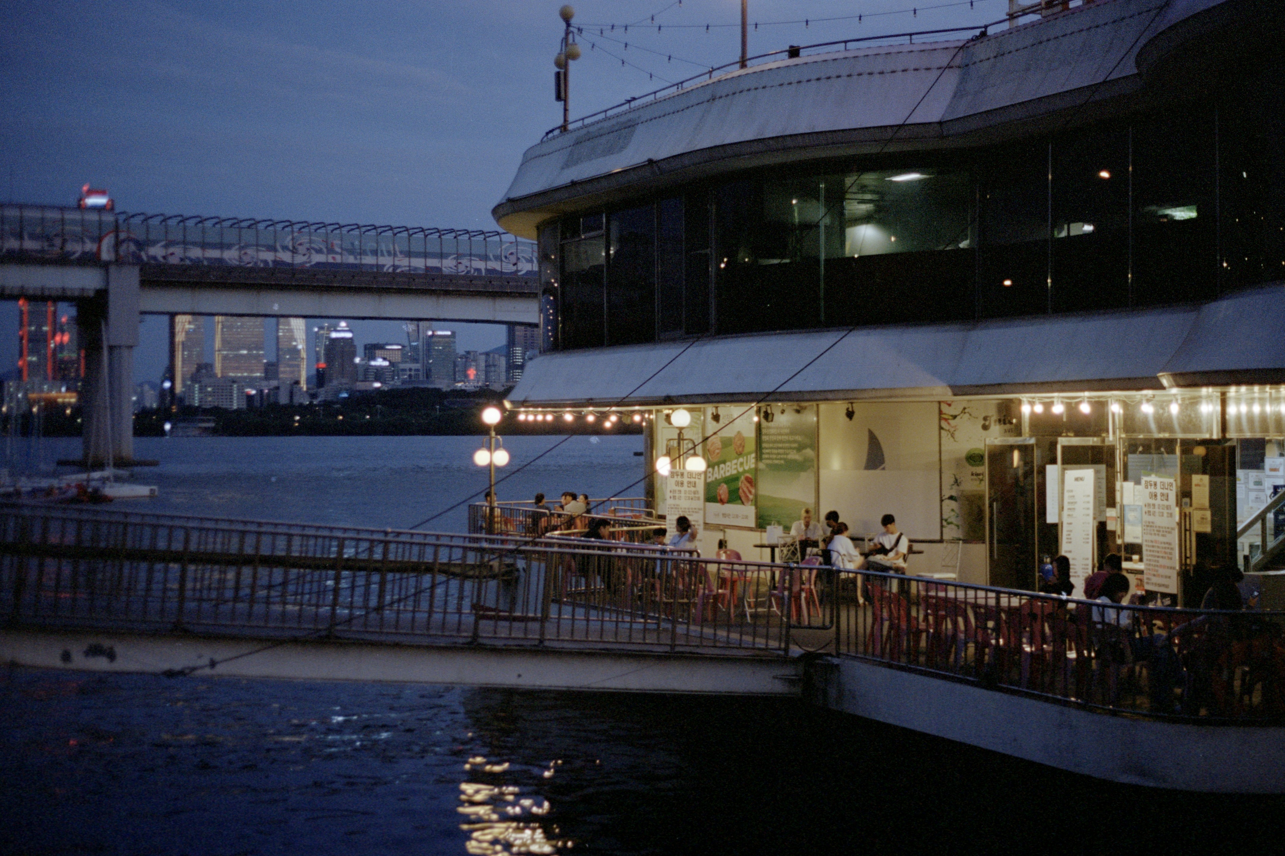 A restaurant on the side of a river with a bridge in the background