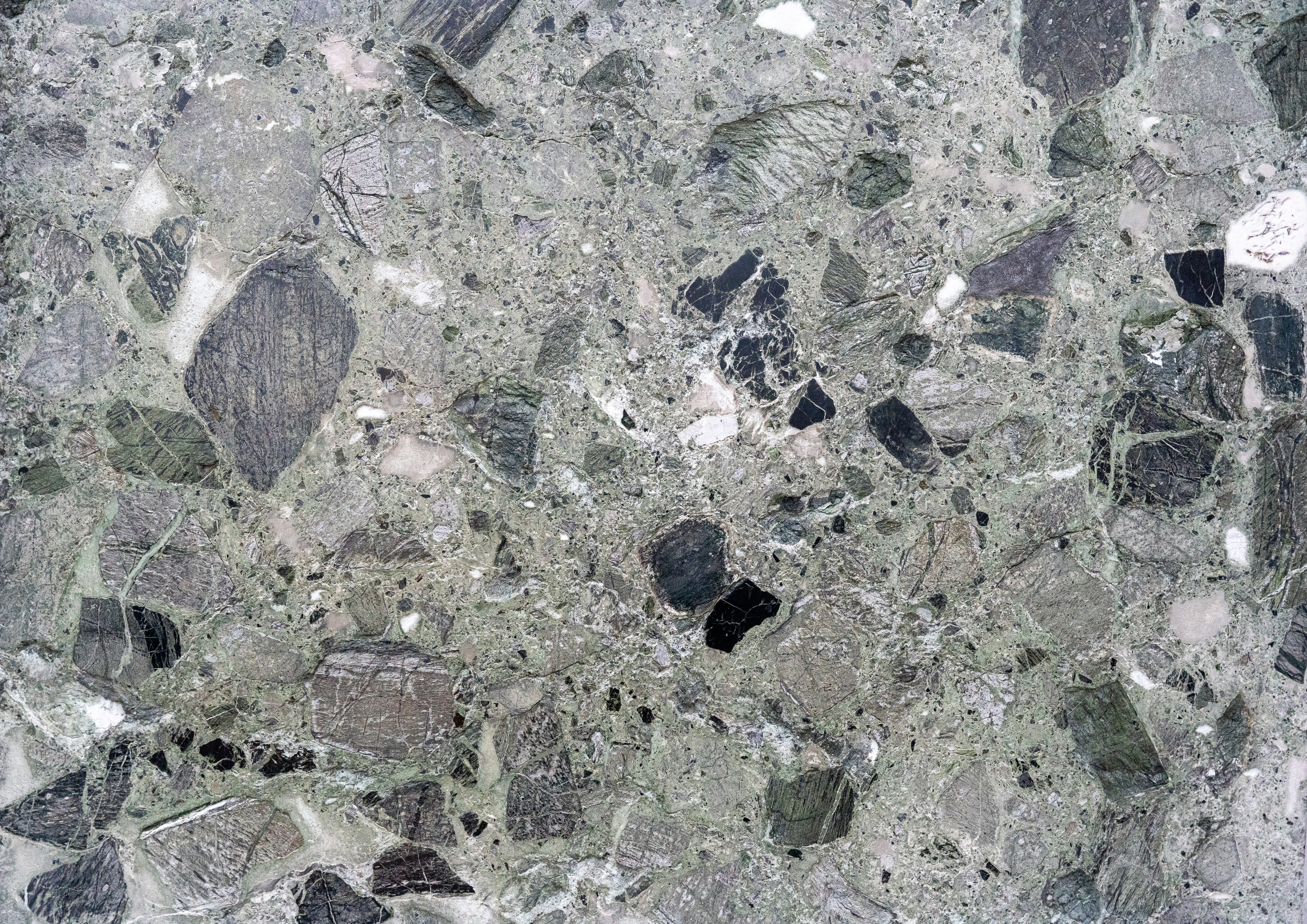 A detailed close-up of a polished stone surface showcasing a mosaic of textured patterns in shades of green, gray, and black. The intricate interplay of angular fragments and earthy tones evokes a raw, natural elegance with a rugged sophistication.