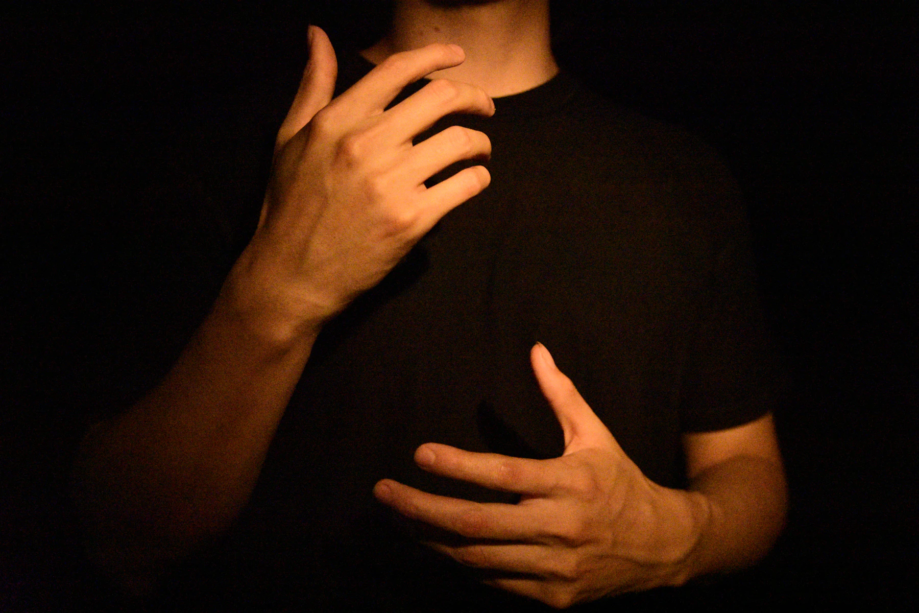 A person in a black shirt signing, one hand raised to the shoulder and the other open at the waist, lit against a dark background