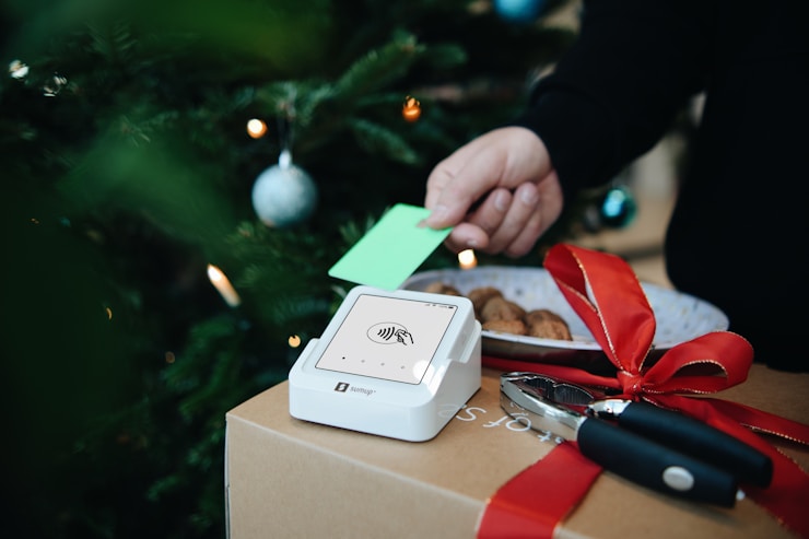 A cozy Christmas scene with a SumUp card reader on a wrapped gift box, surrounded by festive decorations and a green card being used for payment.