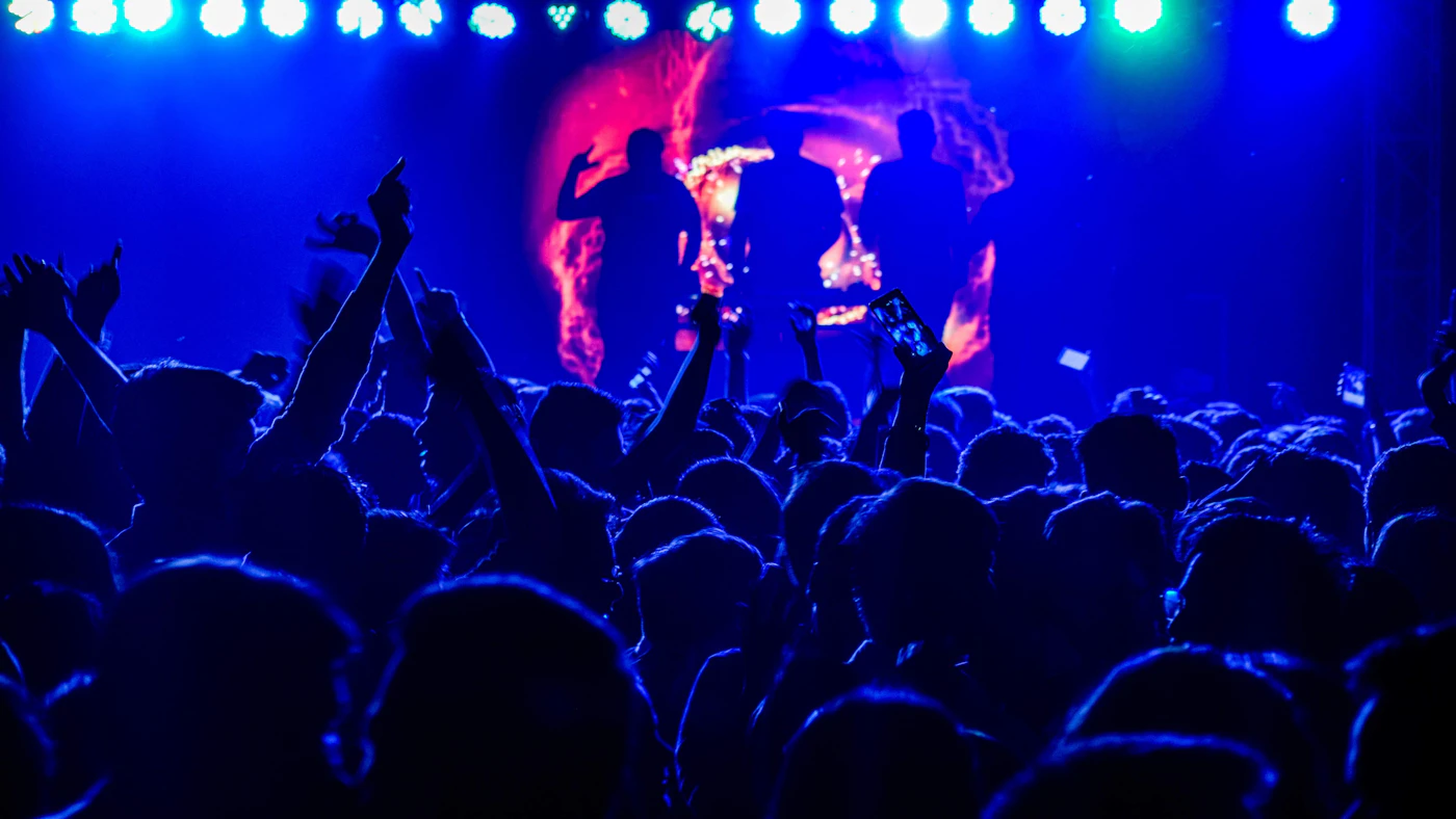 Concert crowd at a venue with arms raised toward the lights