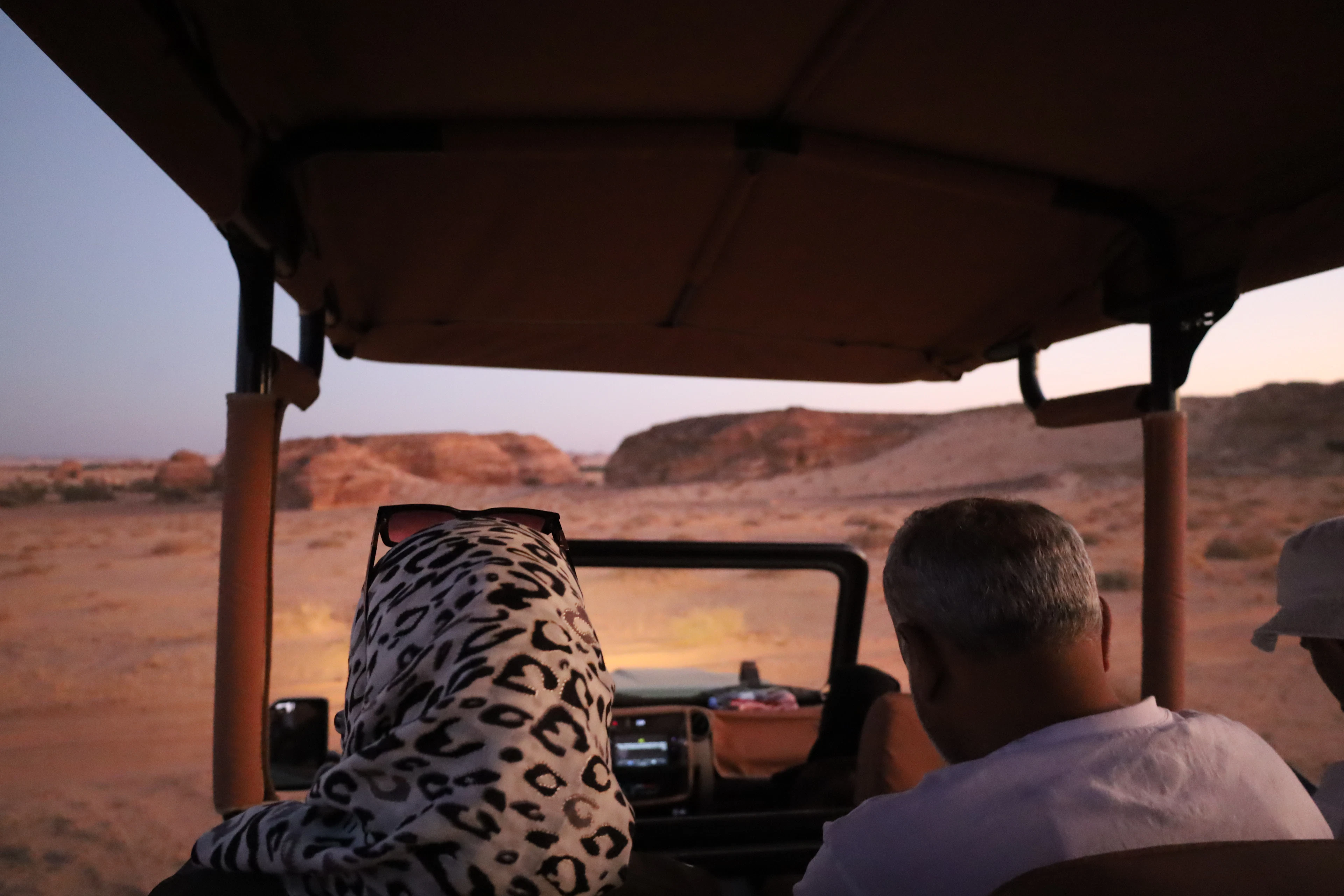 Passengers ride an open-top safari jeep across the desert at dusk, sandstone outcrops glowing in the distance.