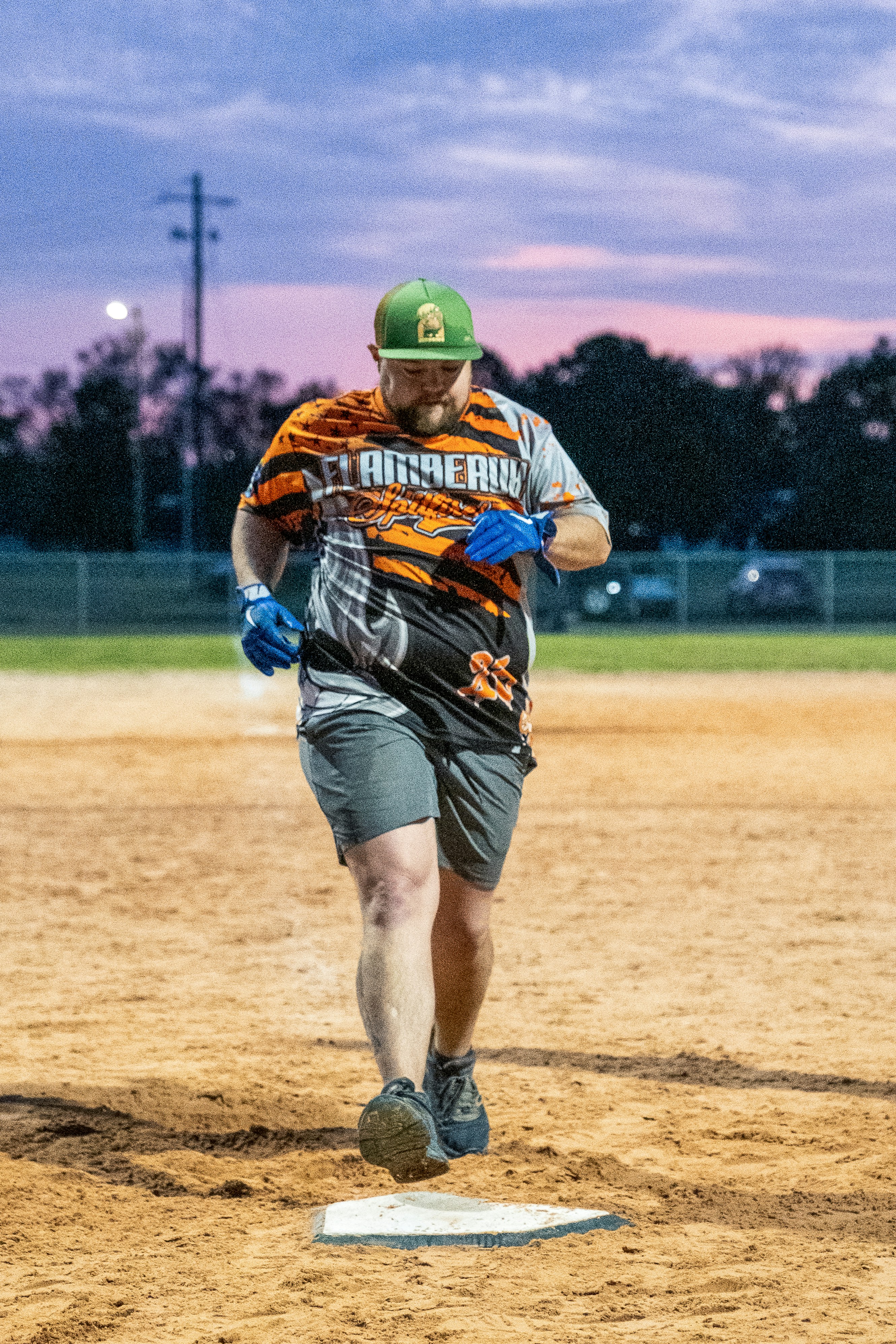 High school softball player running bases at dusk, stadium lights glowing, intense competitive action