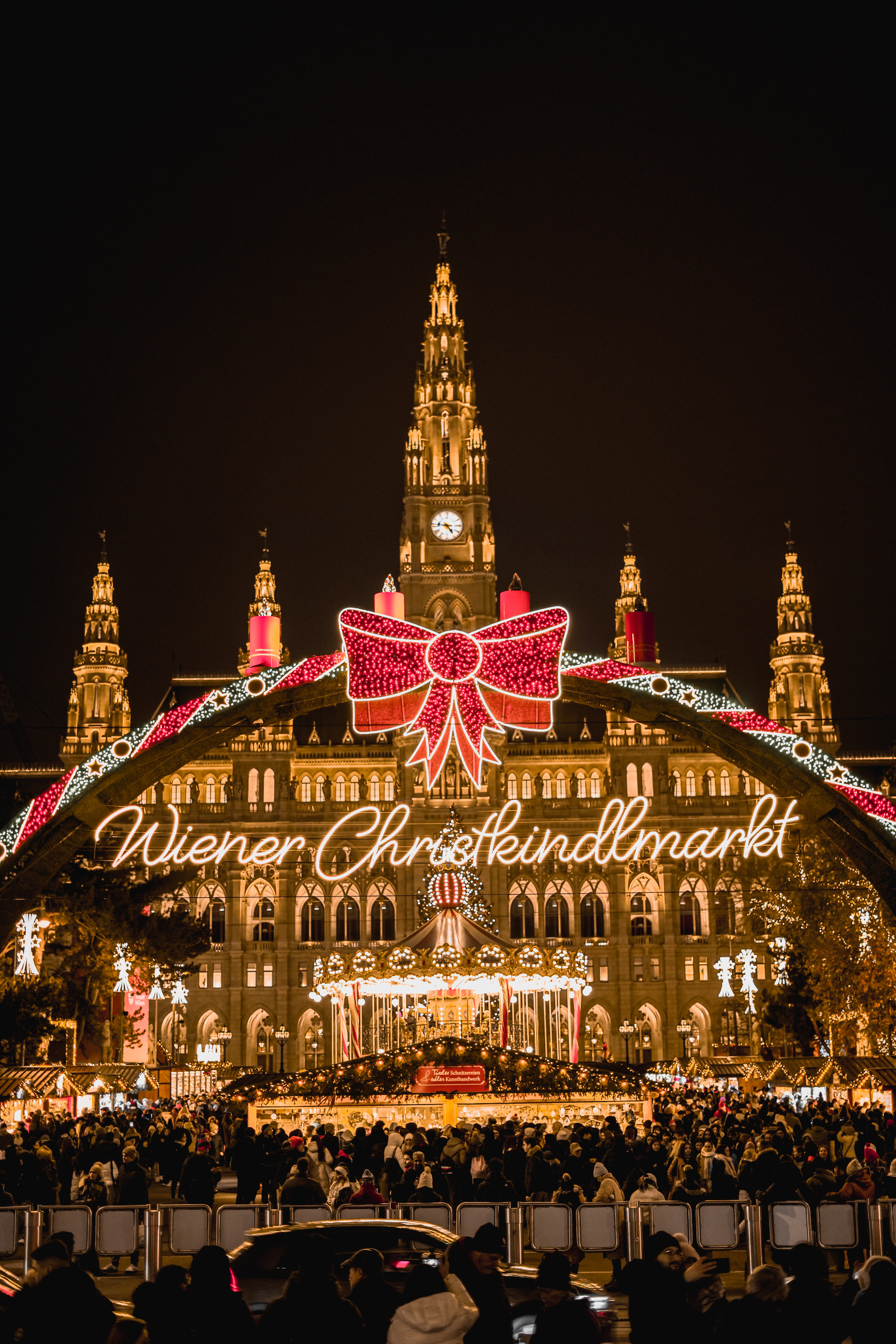 The illuminated arch reading 'Wiener Christkindlmarkt' with its giant red bow at the entrance to Vienna's Christmas market, the neo-Gothic tower of the City Hall lit gold behind it and a crowd filling the square between the lit stalls and the carousel.