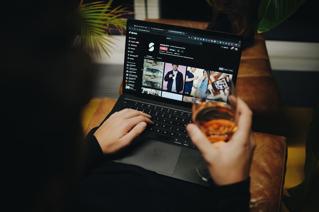 A person sitting on a couch with a laptop, A person holding a glass of wine while browsing TikTok on a laptop, featuring a cozy and modern workspace with green plants in the background.