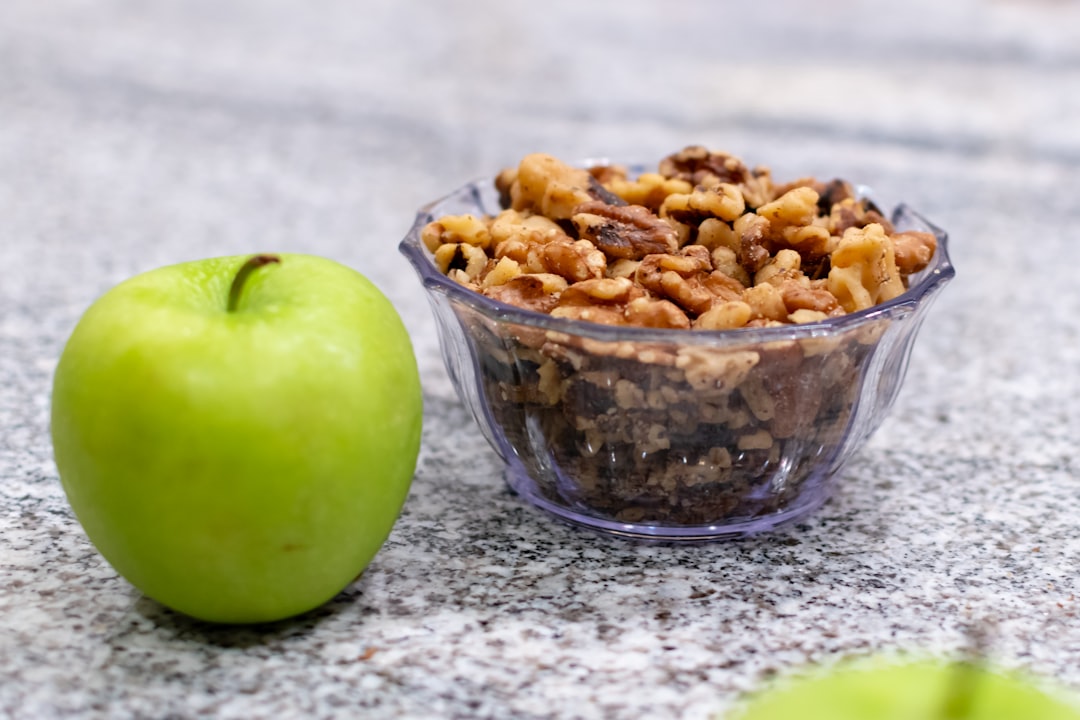 A bowl of granola next to an apple, A green apple sitting beside a bowl of walnuts.