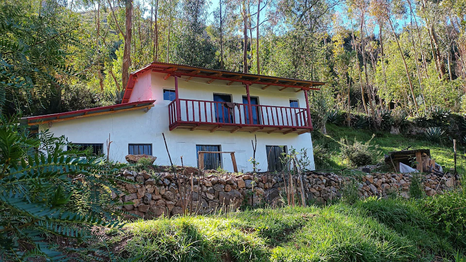 The image depicts a small white house with a red roof nestled amidst lush greenery on a hillside. The house features a balcony, and there are several trees surrounding it, creating a serene and tranquil atmosphere. In the background, more trees can be seen, adding to the natural beauty of the scene.