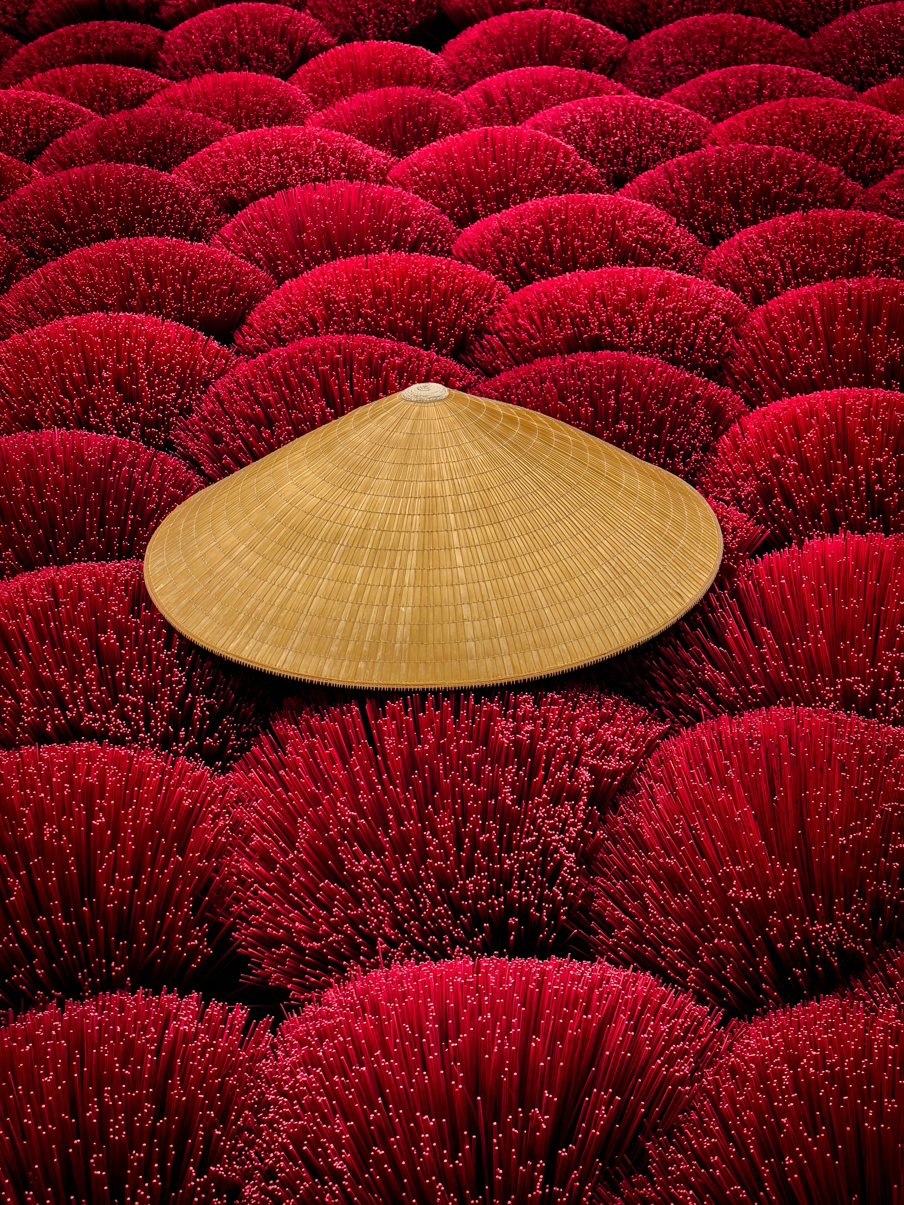 A traditional bamboo hat lies atop bundles of vibrant red incense sticks arranged in circular formations. The scene highlights a cultural craft, with the incense sticks neatly laid out for drying in a symmetrical and visually striking manner.