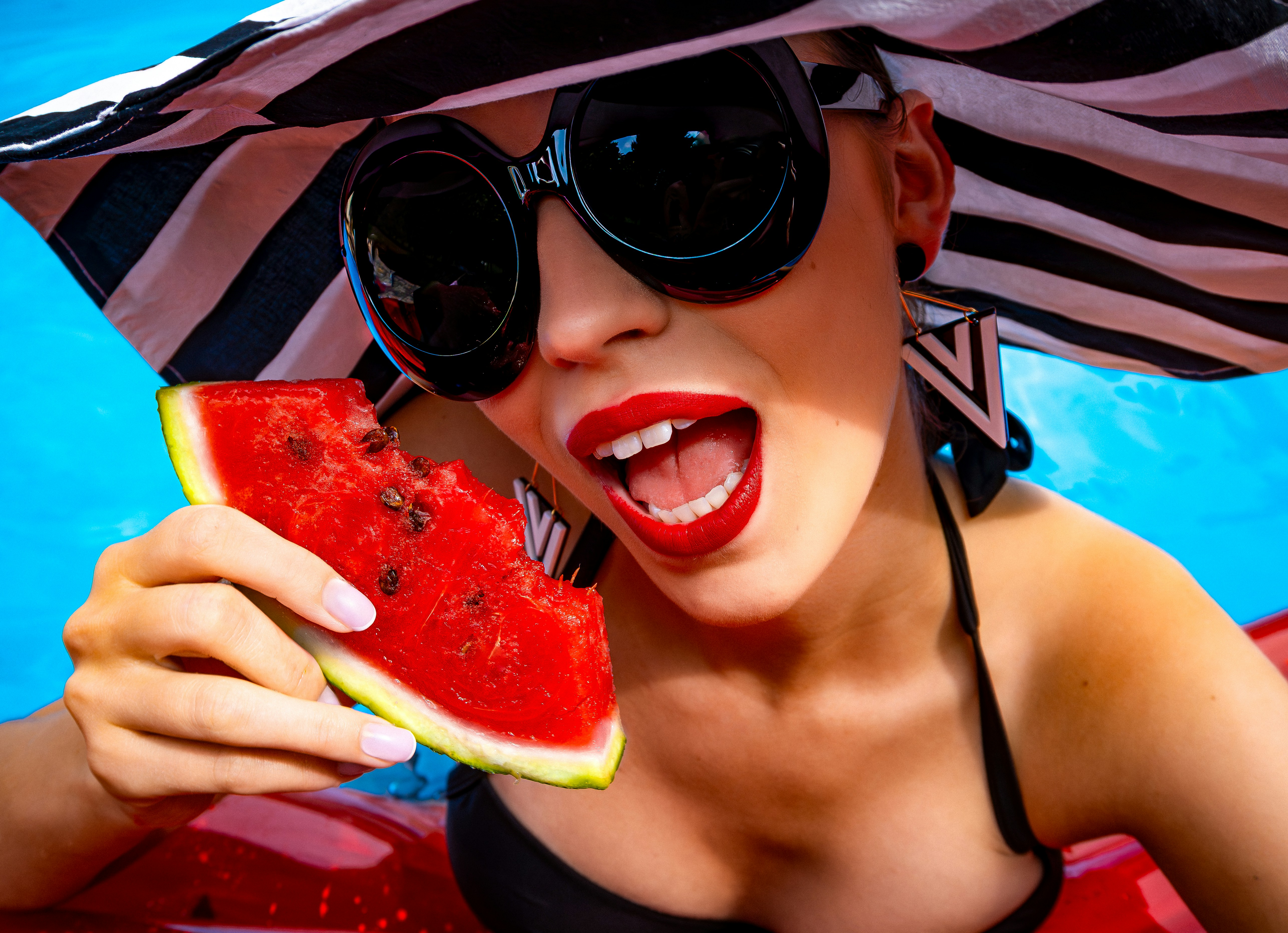 A woman wearing a hat and sunglasses holding a slice of watermelon