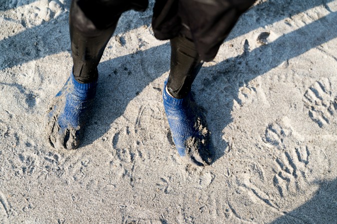 Looking down at feet in sand-caked blue water socks below black wetsuit legs, standing on pale beach sand crossed with footprints