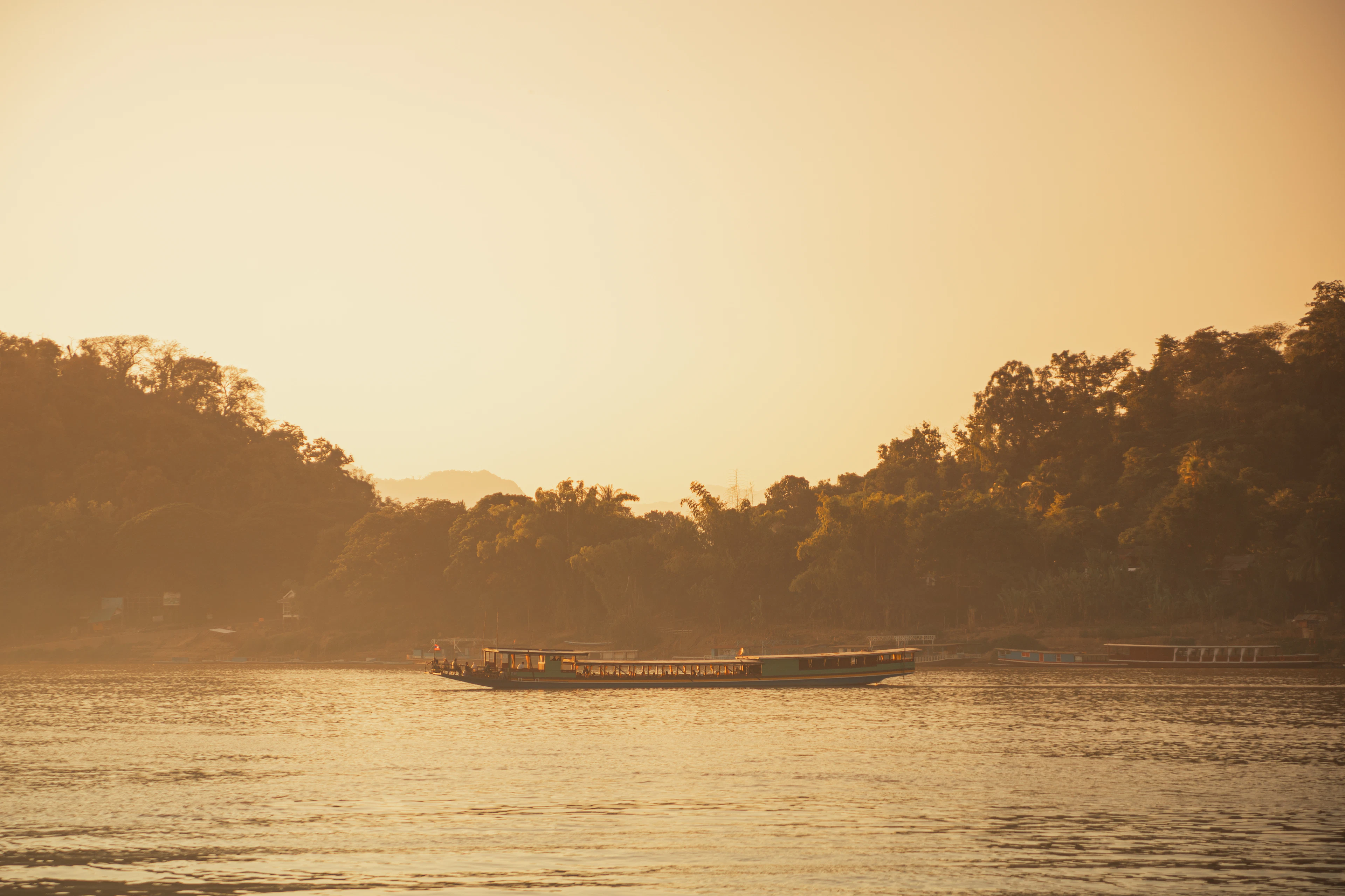 A longtail boat crosses the Mekong River in golden late-afternoon haze, framed by forested banks.