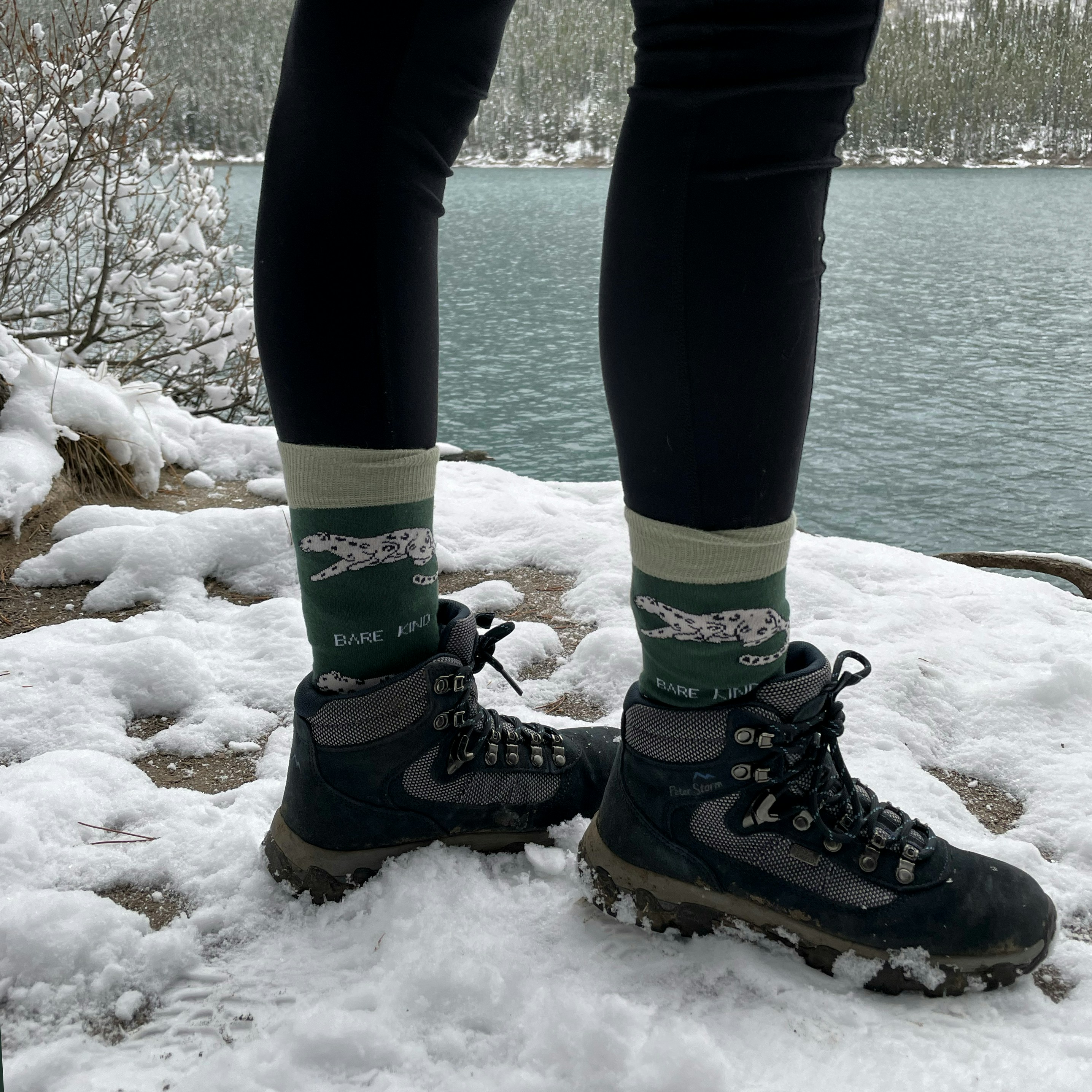 A person standing on snowy terrain beside a lake wearing socks and hiking boots
