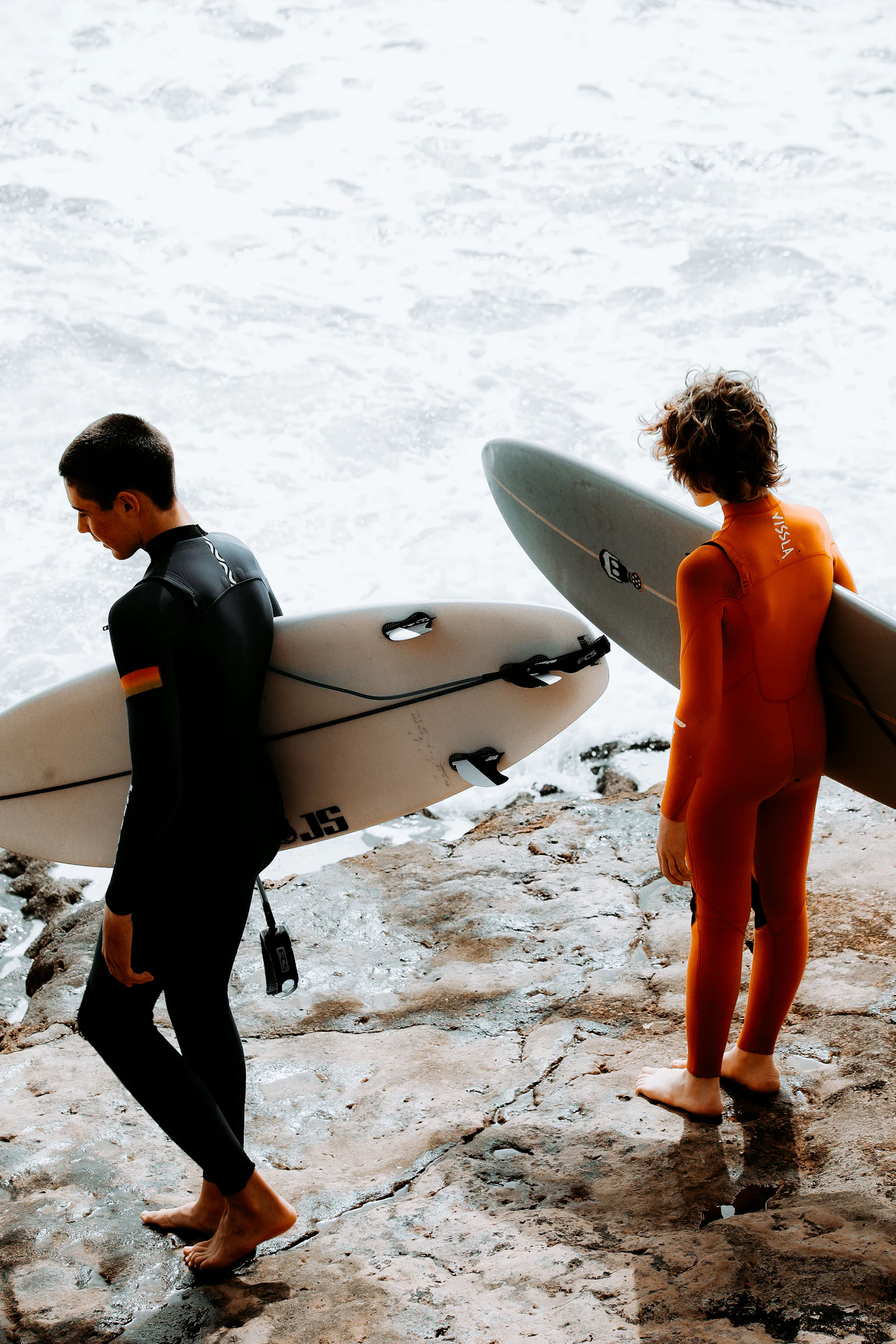 Two surfers carrying boards on the rocky Taghazout shoreline