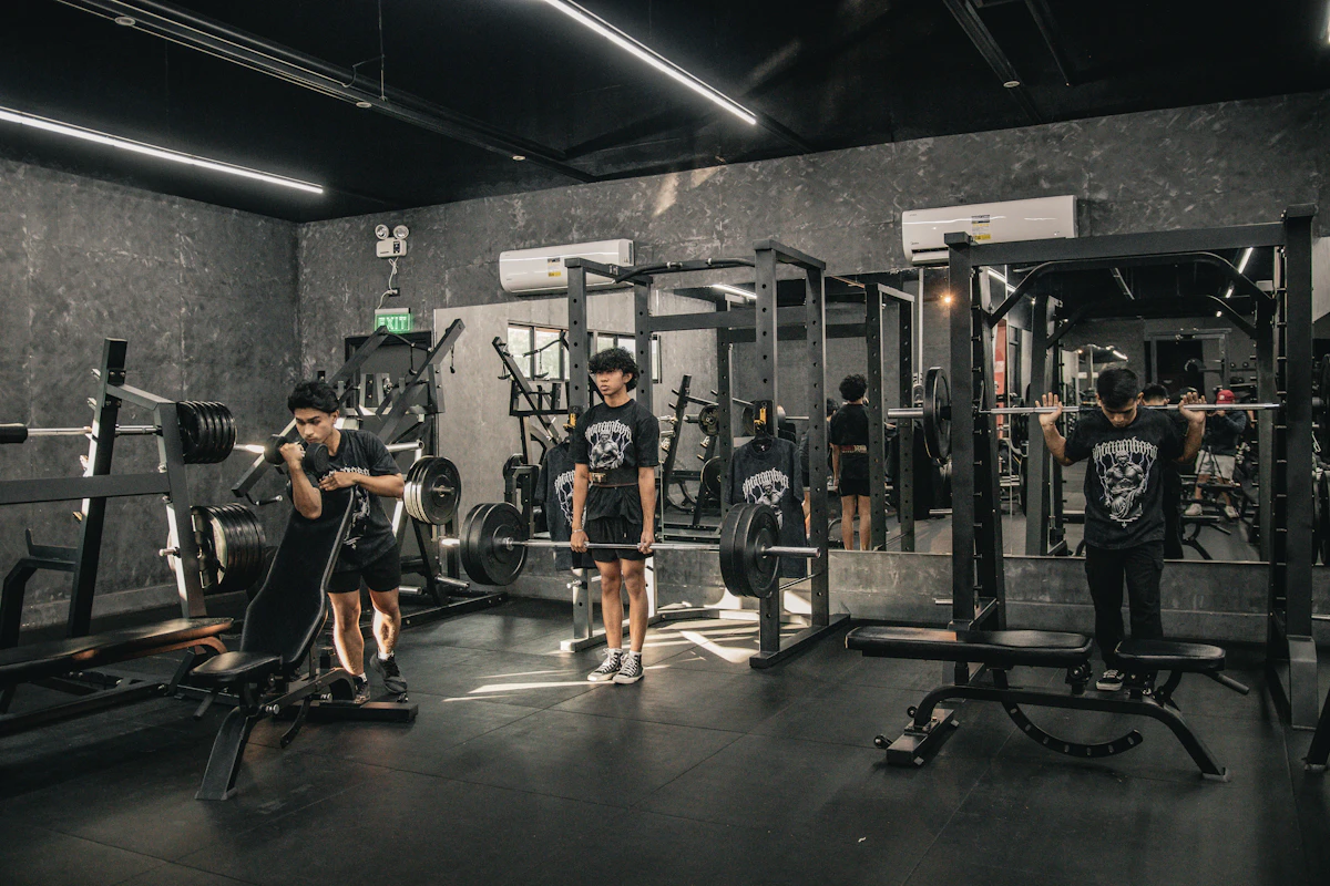 Trois personnes s'entraînent dans une salle de musculation aux murs sombres, entre bancs, racks à squat et disques de fonte