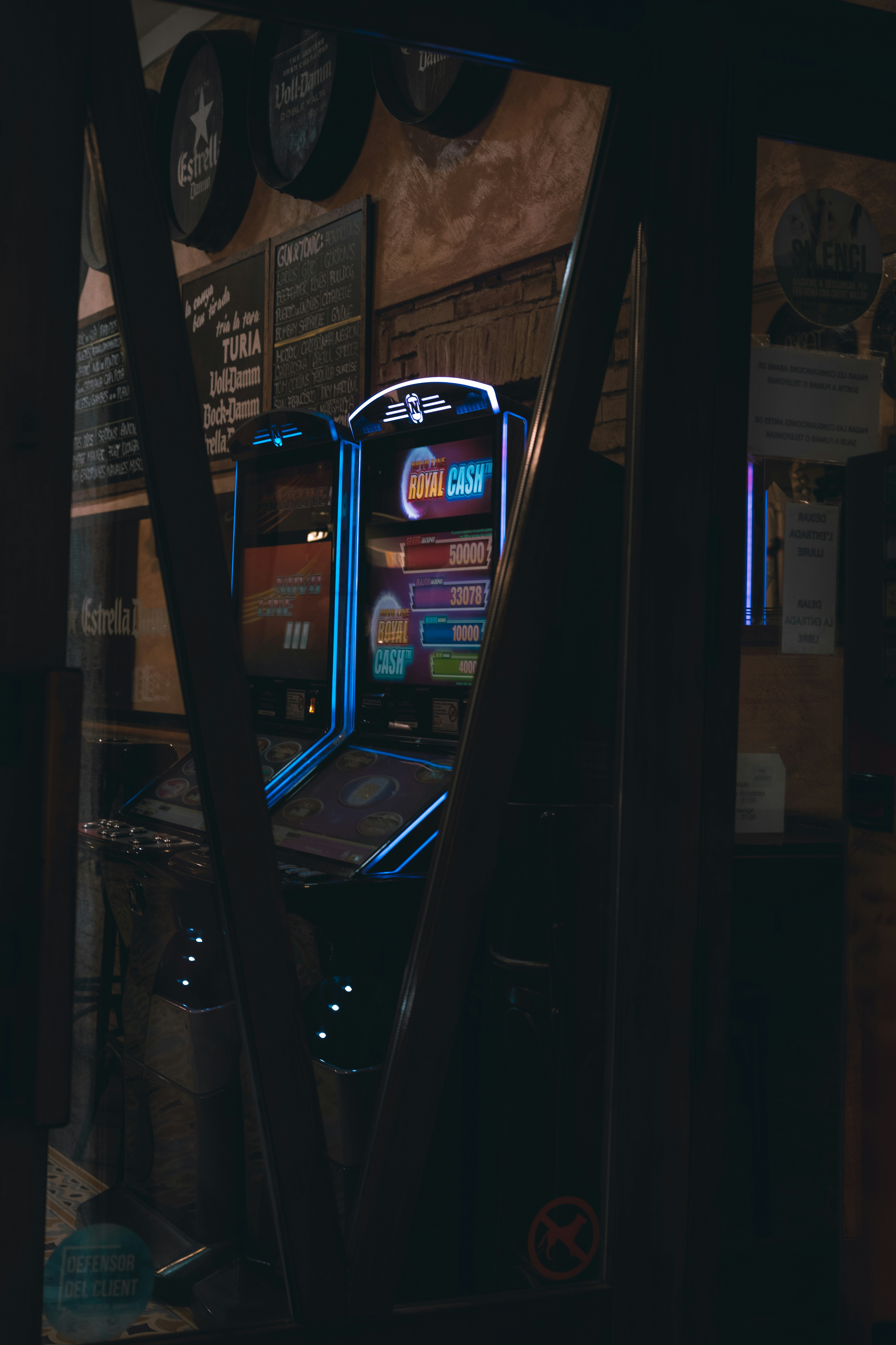 A captivating shot of a slot machine glowing in vibrant neon hues, captured through a reflective glass pane. The dimly lit ambiance of a bar adds depth and contrast to the bright digital display, evoking a sense of nostalgia and intrigue.