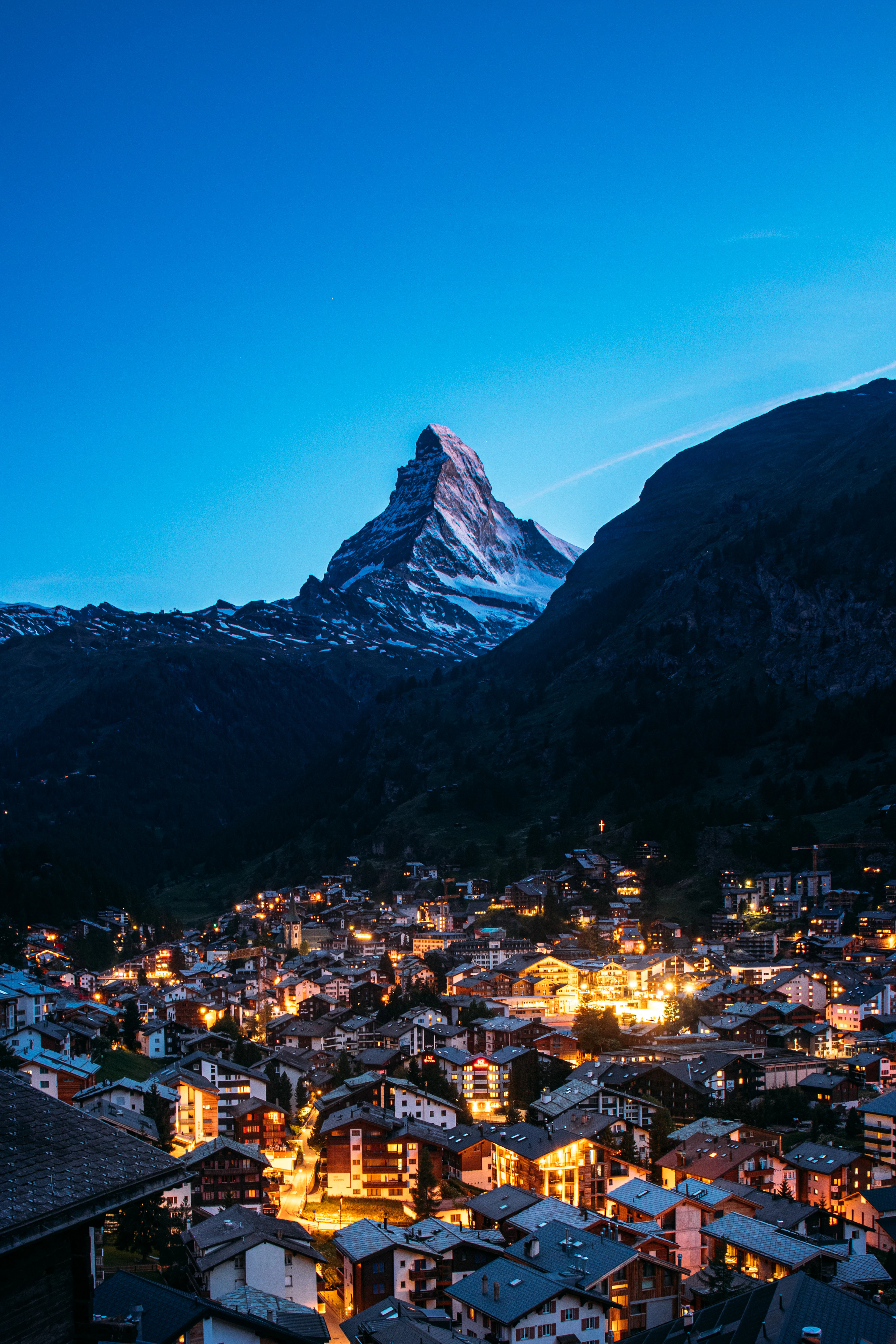 Zermatt's chalets and hotels glowing amber at blue hour, packed along the valley floor beneath the pale snow-streaked pyramid of the Matterhorn under a deep cobalt sky