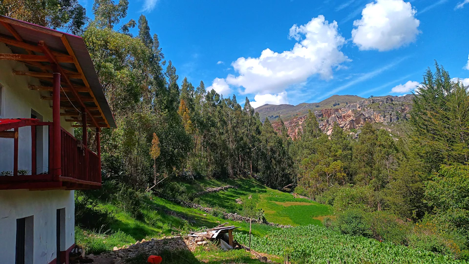 The image depicts a serene mountainous landscape with a white house nestled at the foot of a mountain range, surrounded by lush greenery and trees. The house features a red roof that contrasts with the verdant surroundings. A balcony overlooks the picturesque scene from the second floor. In the foreground, there is a small garden with various plants and flowers, adding to the natural beauty of the landscape.