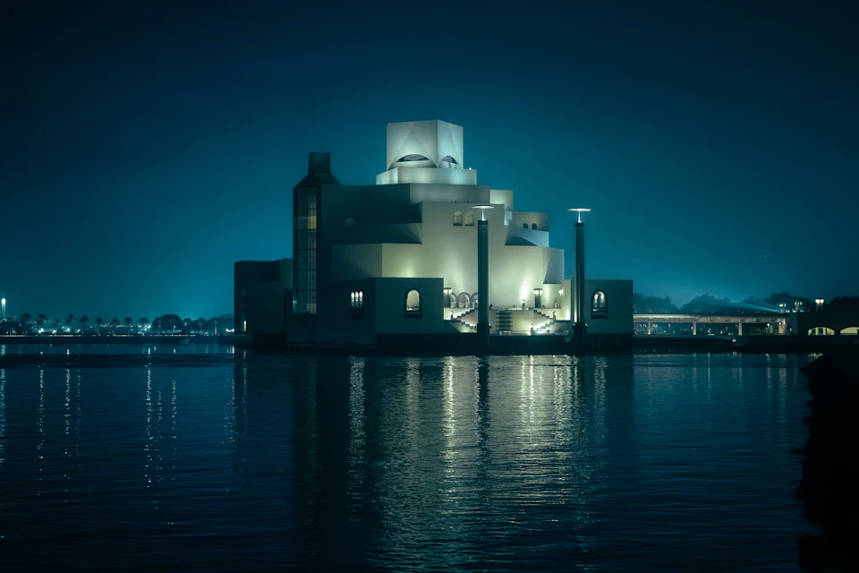 The image depicts a large, white building with a distinctive dome and columns situated on the shoreline of a body of water. The building is illuminated by multiple lights, creating a striking contrast against the dark sky. The perspective of the photo is taken from across the water, providing a comprehensive view of the structure's architectural features.