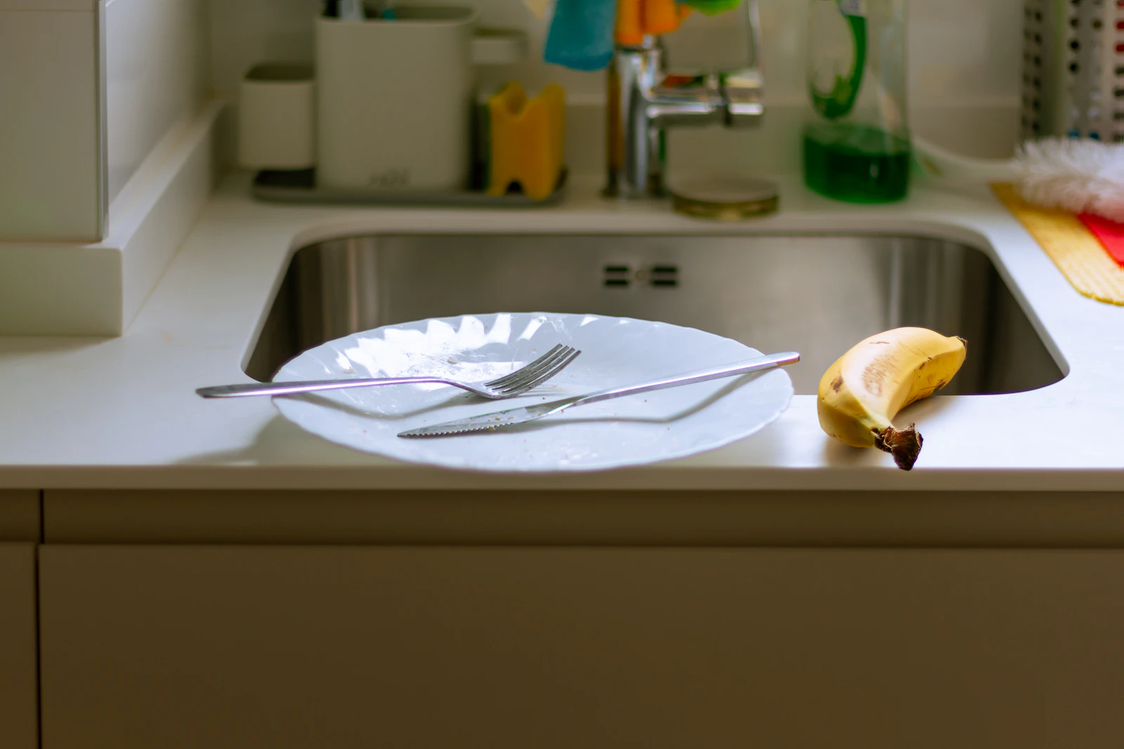 An empty plate with a fork and knife resting by a kitchen sink, a banana skin on the worktop beside it