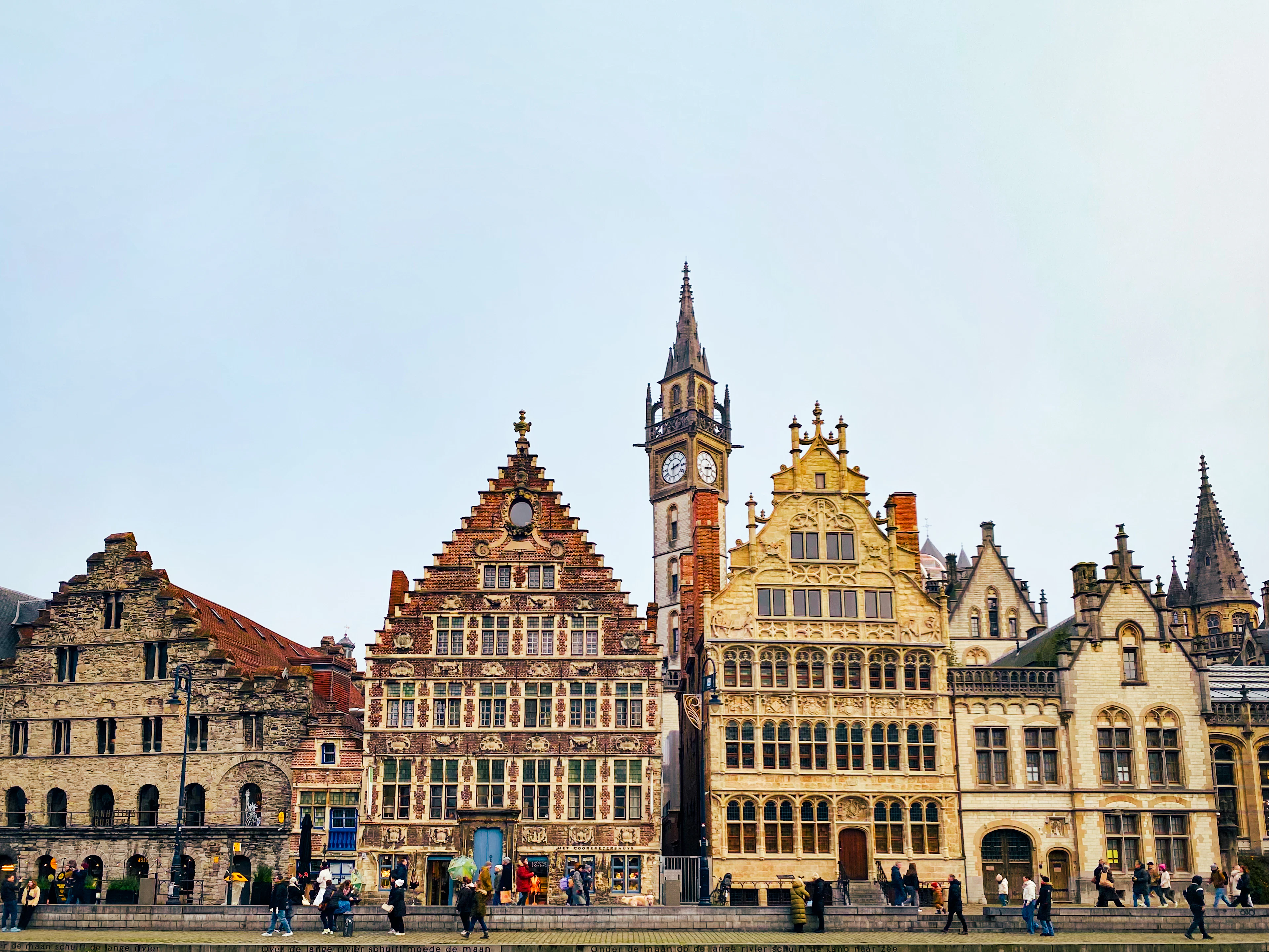 Sunlit stepped gabled guild houses along Ghent's Graslei canal, with the Belfry tower behind and people strolling below.