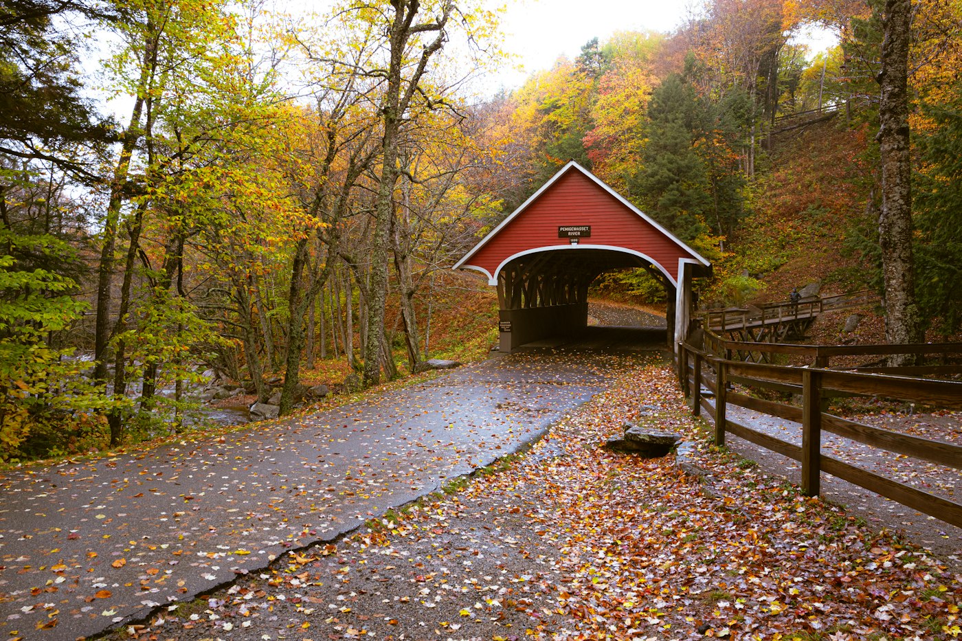 The red Flume Covered Bridge in Lincoln framed by autumn trees and a leaf-covered path, a classic stop on a New Hampshire covered bridges road trip
