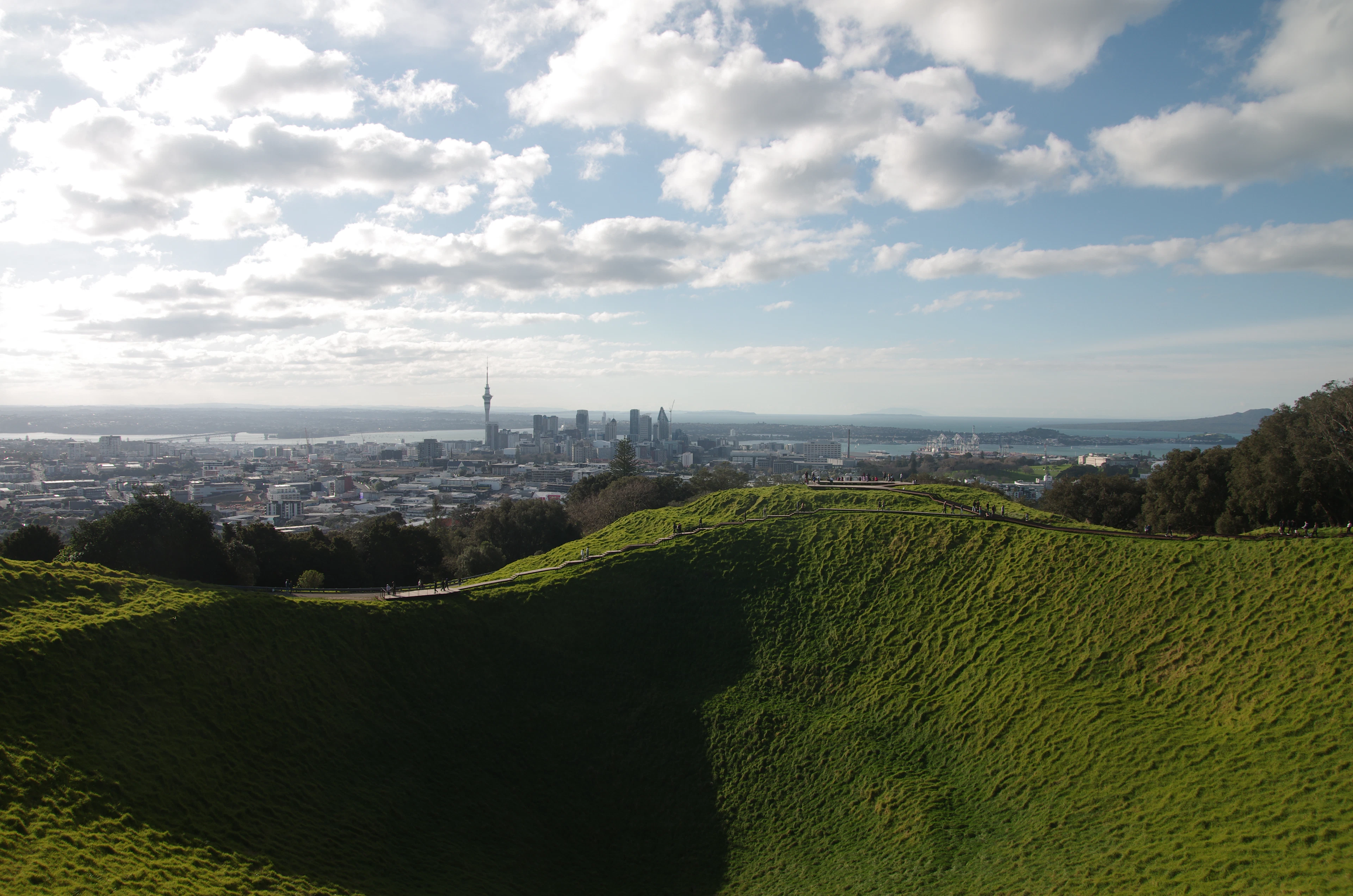 The deep grassy crater of Mount Eden in Auckland catching low sun, with the Sky Tower and the city skyline strung along the harbour beyond